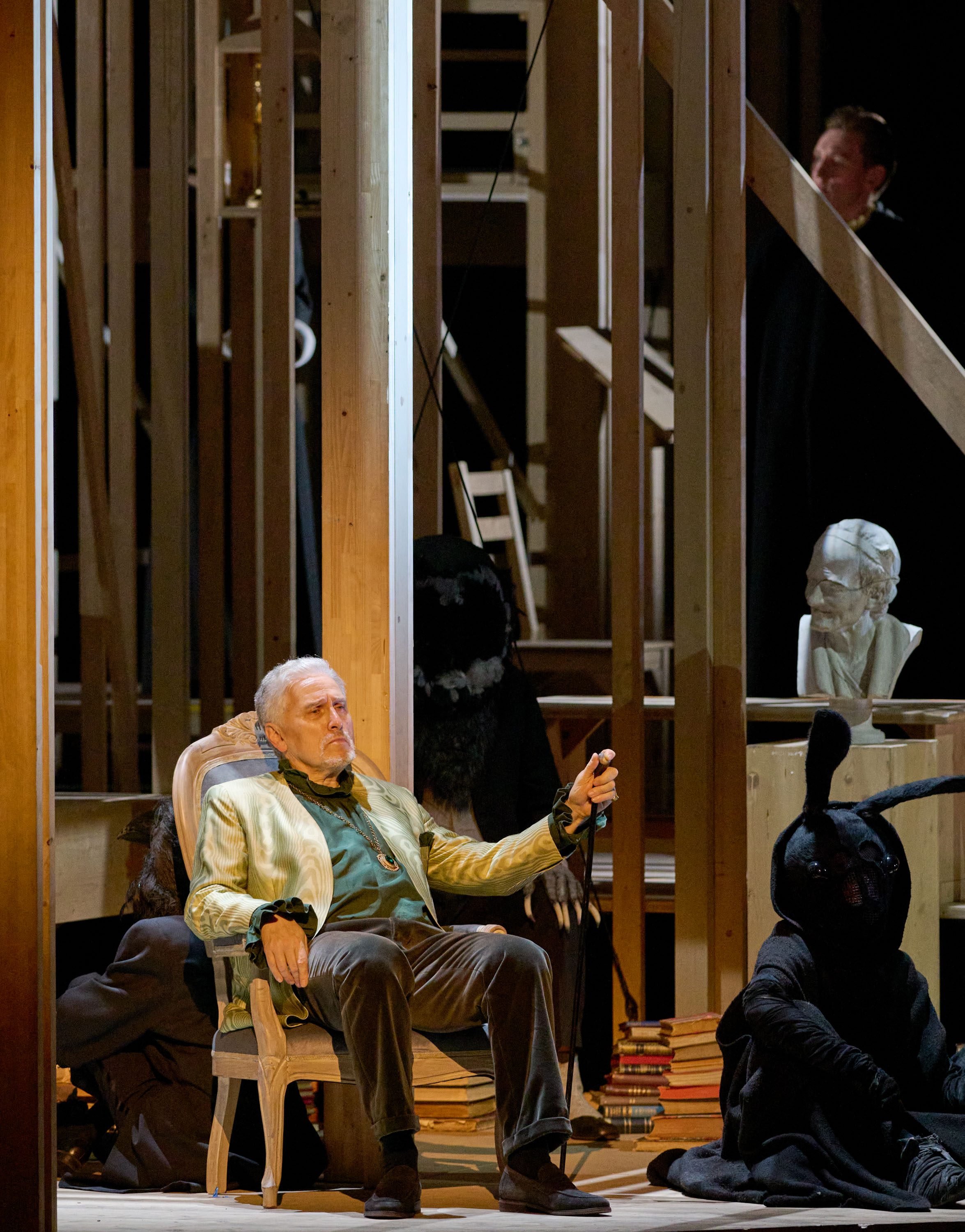 A man in a green and gold jacket sits in a chair, appearing contemplative. Beside him is a figure in a black costume with a hood and bunny ears. The background features wooden scaffolding, books, and a bust statue, creating a theatrical setting.