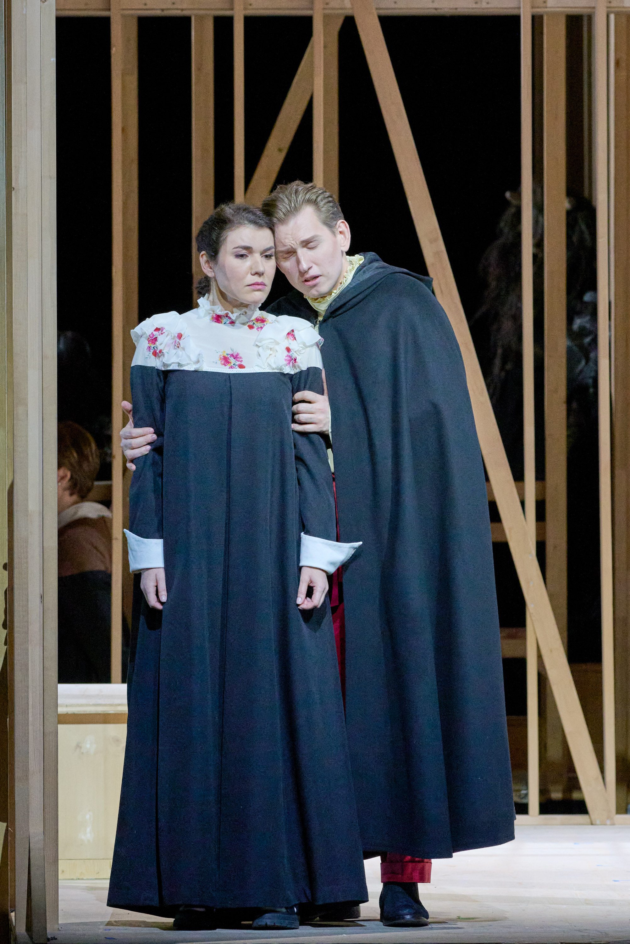 A young man in a dark cape embraces a woman in a long, dark dress with a floral collar. Both appear concerned as they stand together on a stage with a wooden backdrop, suggesting a moment of tension or drama in a performance.