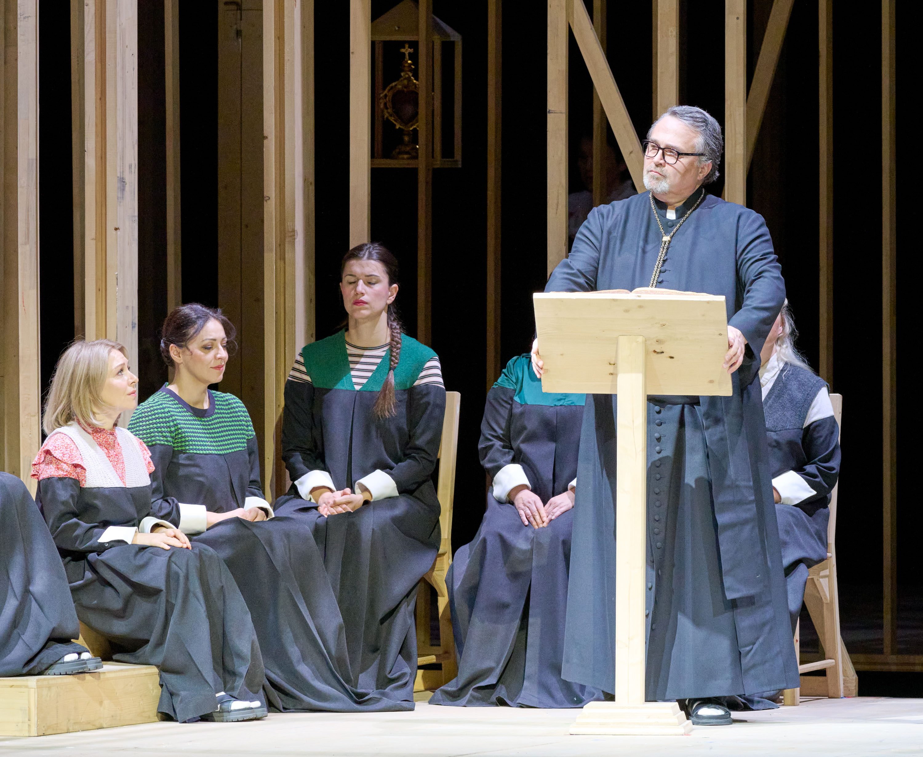 A group of people, dressed in dark robes, sits on a wooden stage while a man in a priest's attire stands at a podium, delivering a speech. The audience appears attentive, with a mix of expressions among the seated individuals. The backdrop consists of wooden structures.