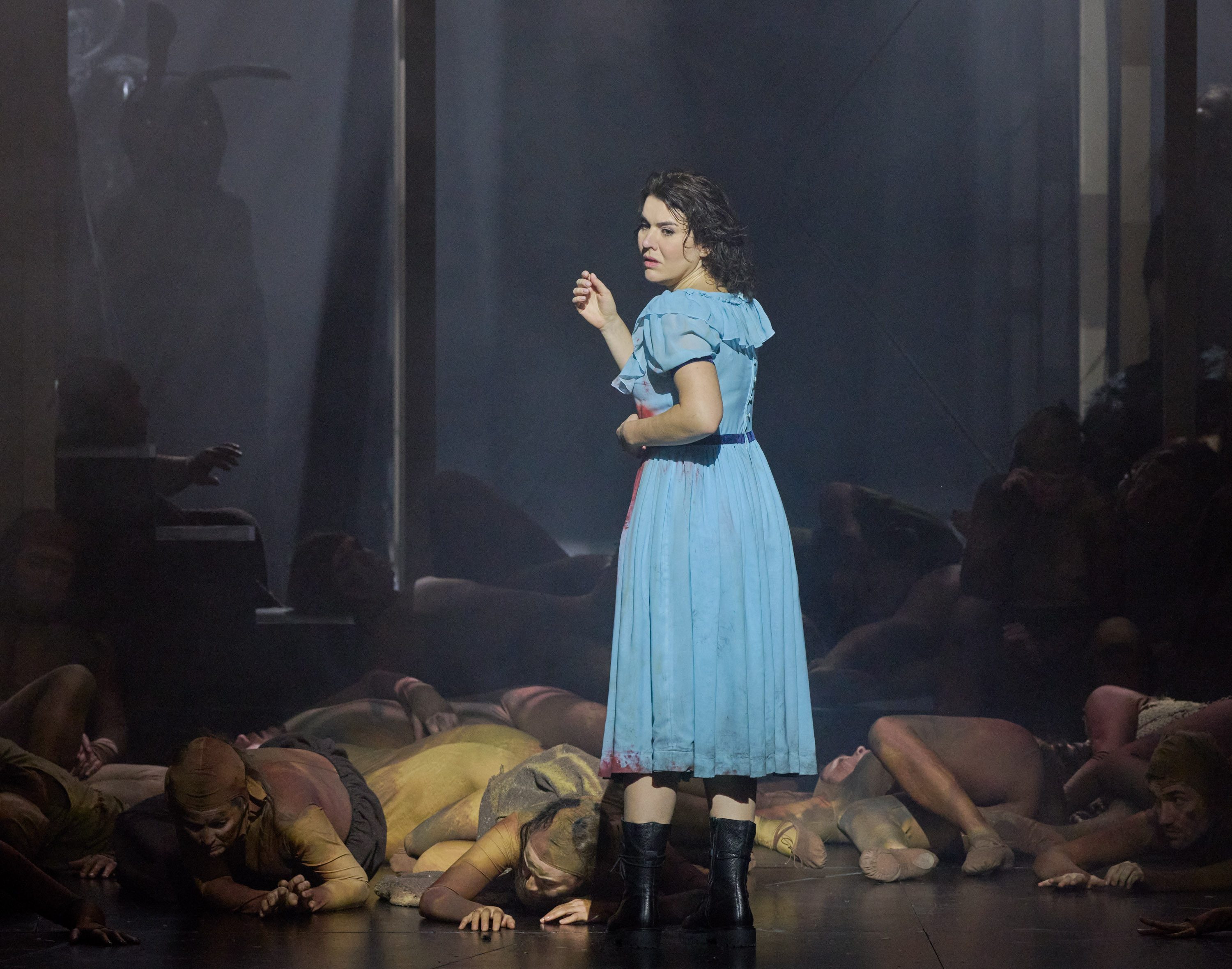 A woman in a blue dress stands on stage, looking concerned. She is surrounded by people lying on the floor, suggesting a dramatic scene. The atmosphere is dimly lit with soft fog, creating a tense and emotional environment.