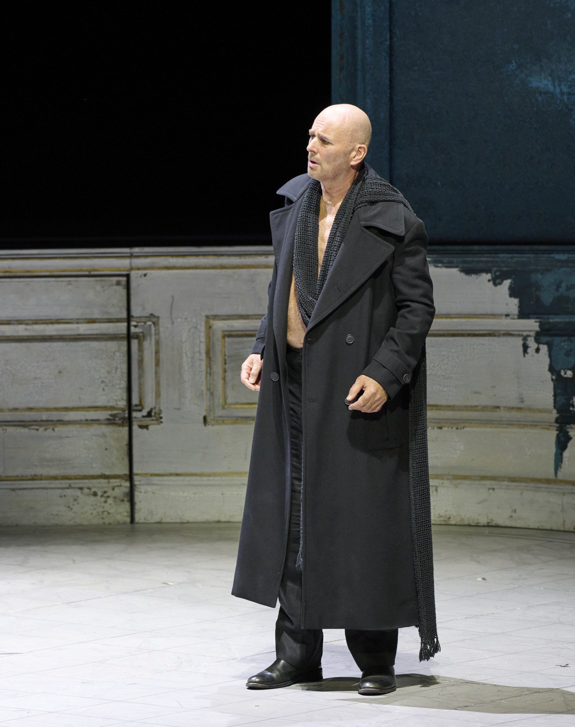 A bald man stands on stage wearing a long black coat and a scarf, his shirt unbuttoned. He appears to be in a dramatic pose, possibly delivering a passionate speech or performance. The background is minimal and dark, focusing attention on the figure.