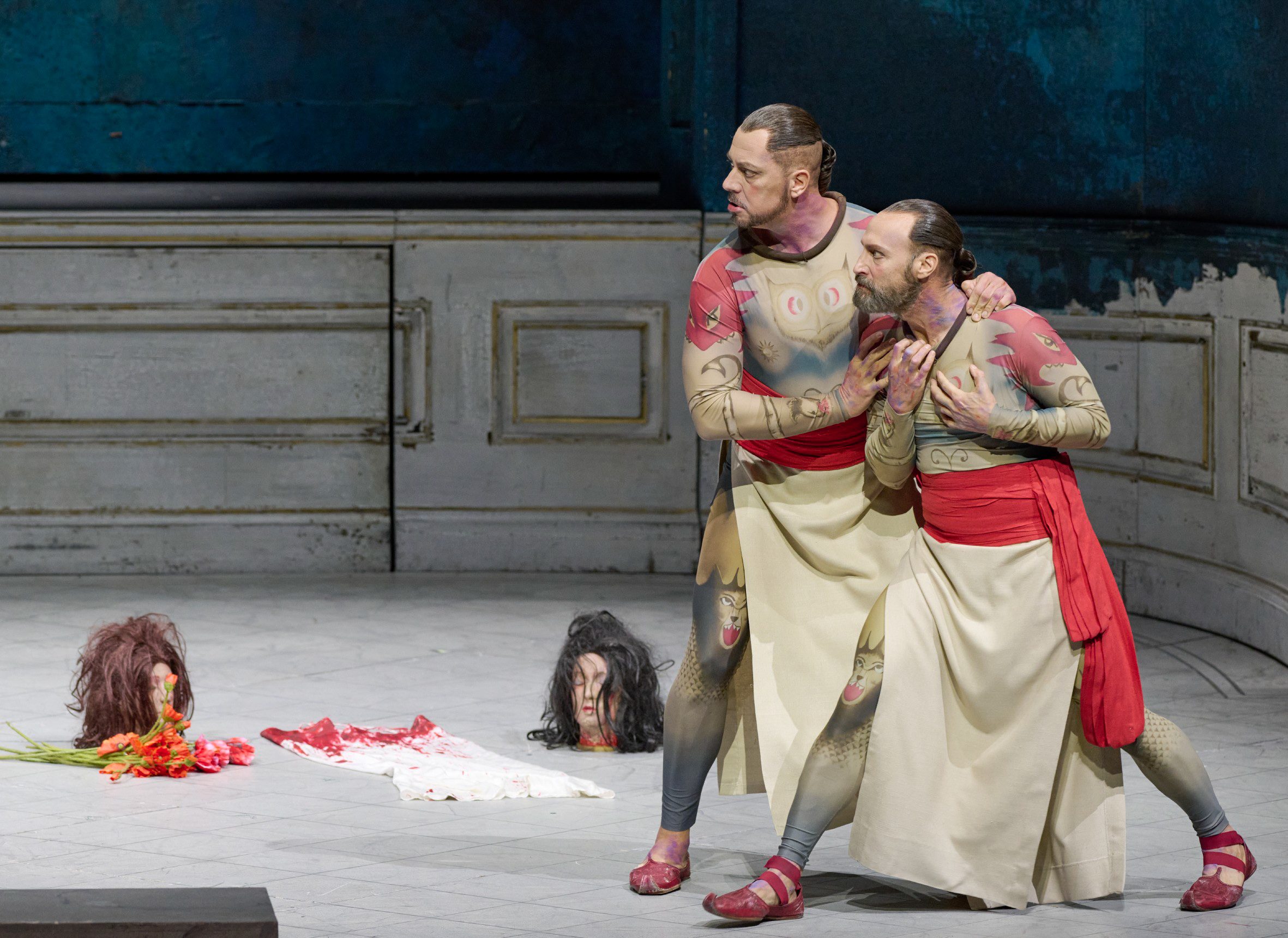 Two male actors in stylized costumes are stage acting, appearing distressed as they embrace each other. In the foreground, two severed heads and a blood-stained cloth lie on the ground, adding a dramatic element to the performance. The backdrop features textured blue walls.