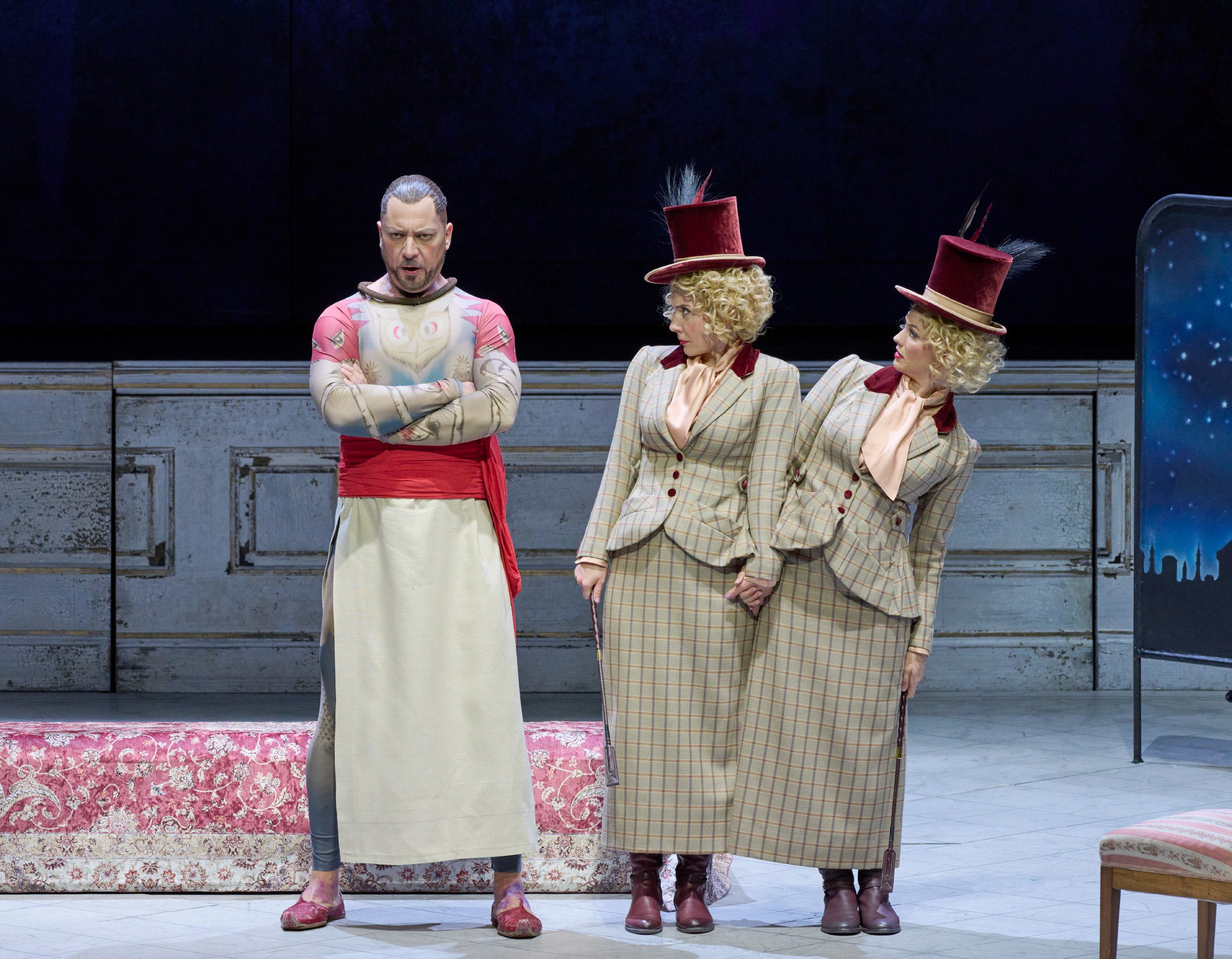 A man in a costume with a cape and intricate body painting stands with his arms crossed, looking stern. Two women, dressed in matching plaid suits and top hats, lean toward each other, appearing curious or playful. They are on a theatrical stage with a simple backdrop.