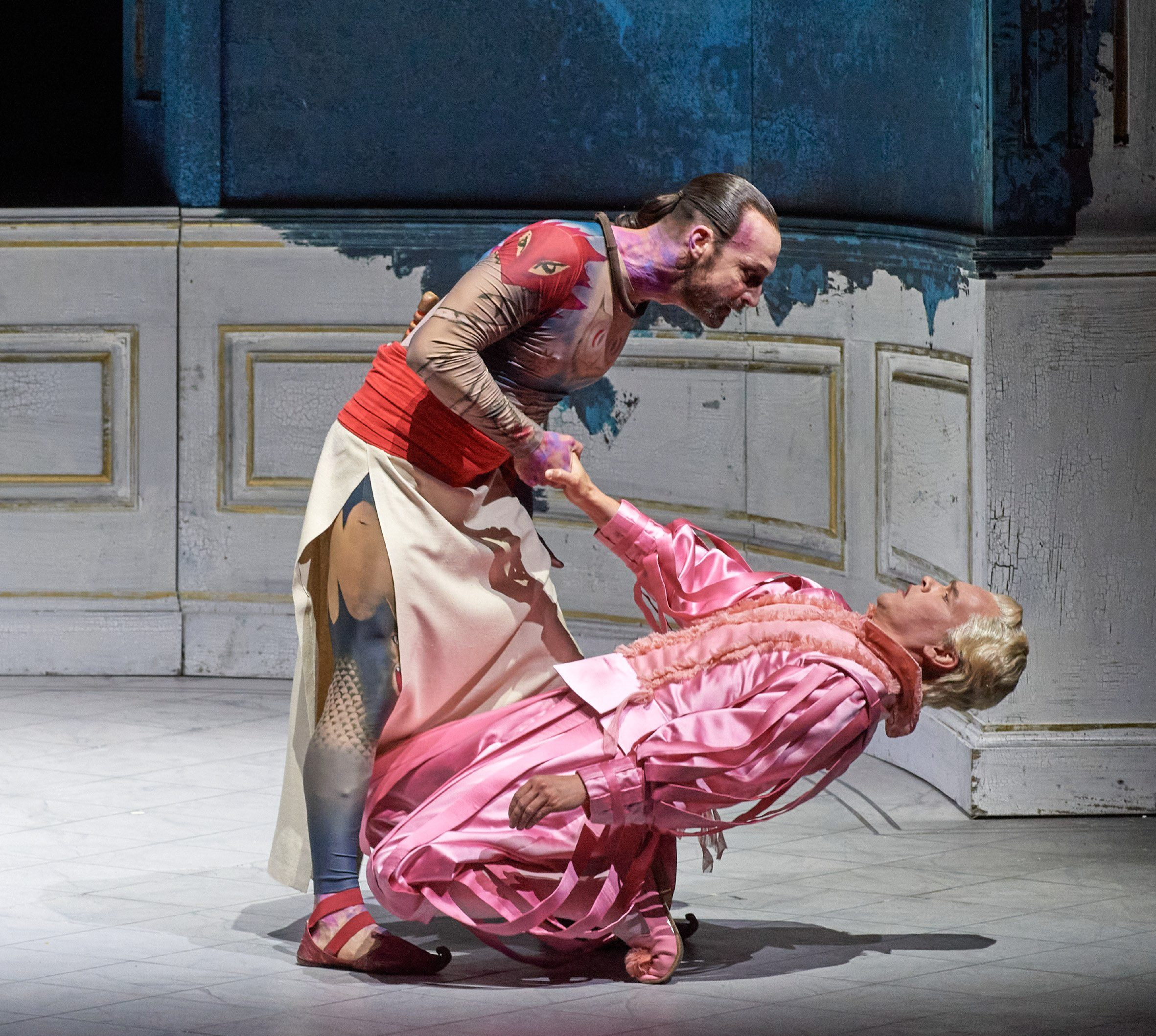 Two male dancers perform on stage. One, in a colorful outfit with a fitted top and flowing skirt, is reaching out to the other, who is dressed in a pink outfit and appears to be falling or leaning back. The backdrop suggests a theatrical setting.