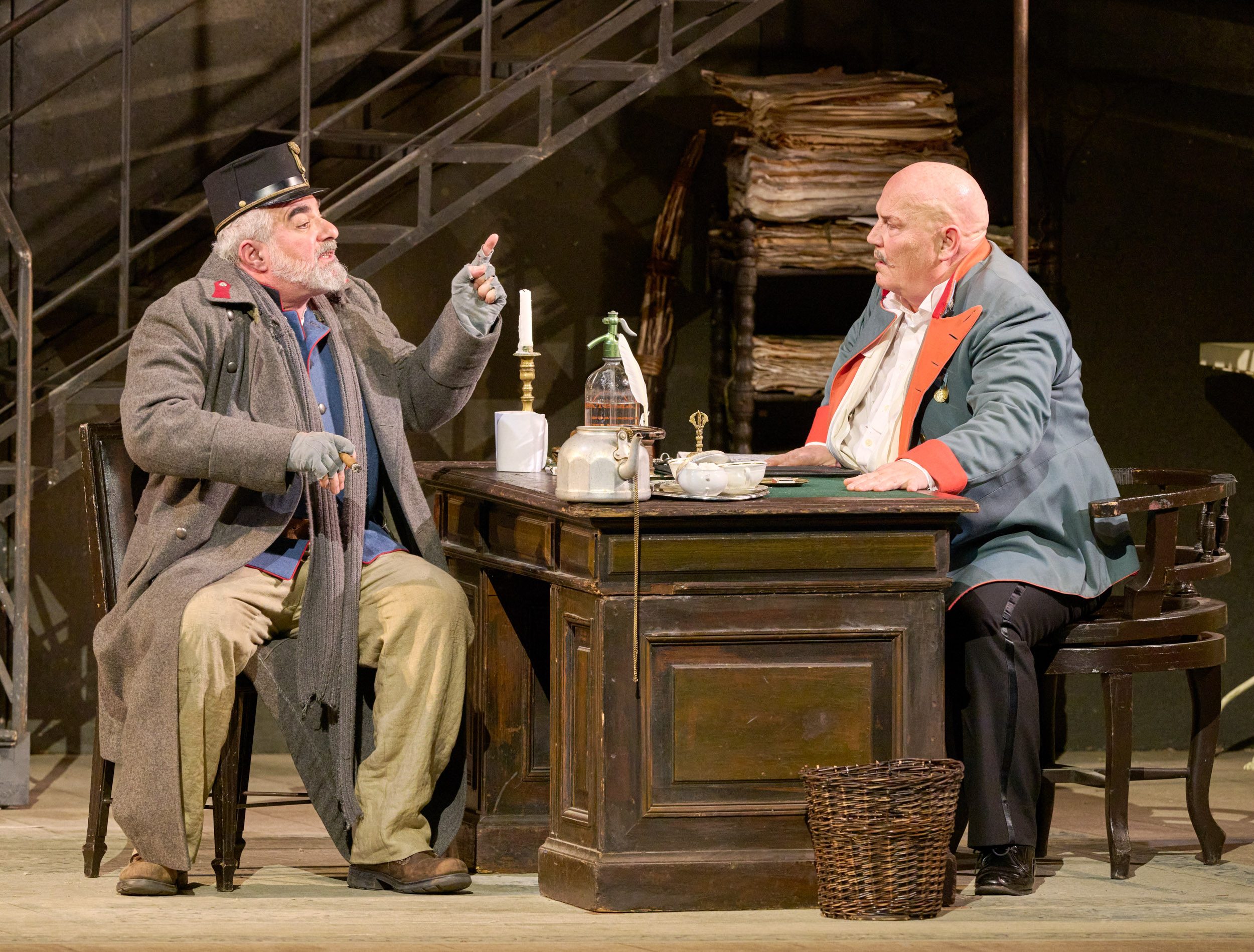 Zwei ältere Männer sitzen sich an einem vollgestellten Schreibtisch gegenüber; einer gestikuliert lebhaft, der andere hört aufmerksam zu.