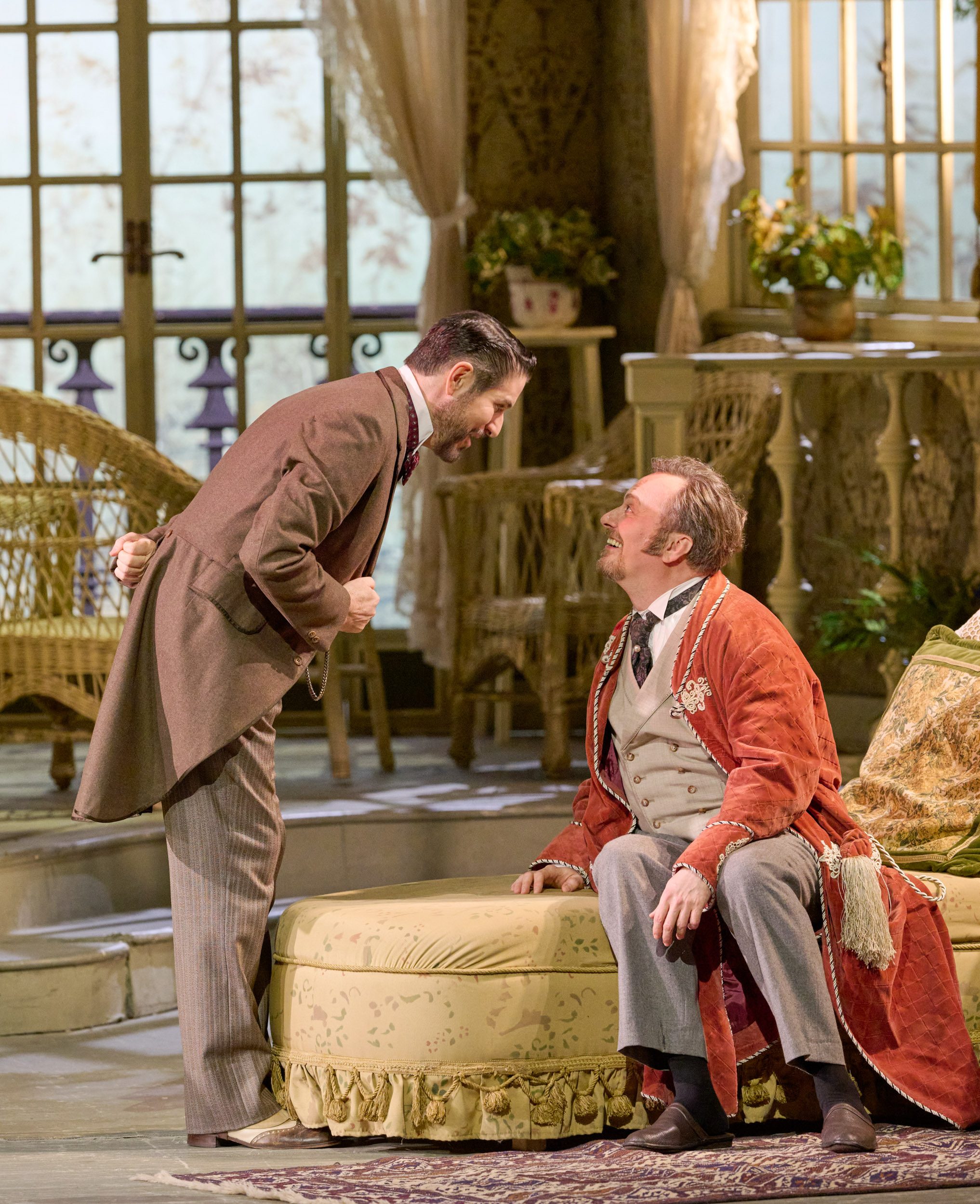 Ein Mann in braunem Anzug beugt sich vor und spricht eindringlich mit einem älteren Mann im roten Morgenmantel, der auf einem Sofa sitzt.