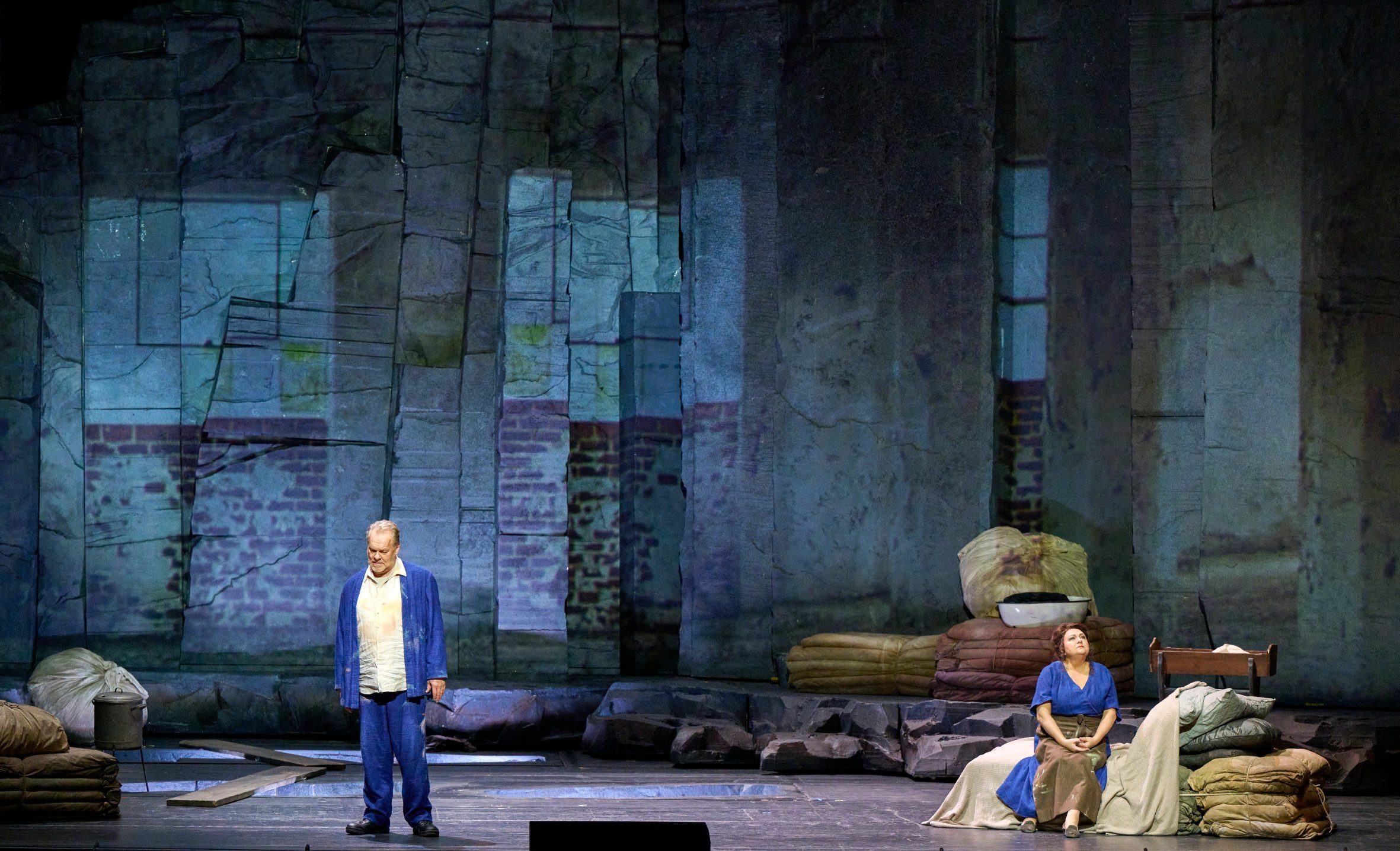 Ein Mann in blauem Outfit steht, während eine Frau in blauem Kleid auf der Bühne vor zerfallenen Mauern sitzt.