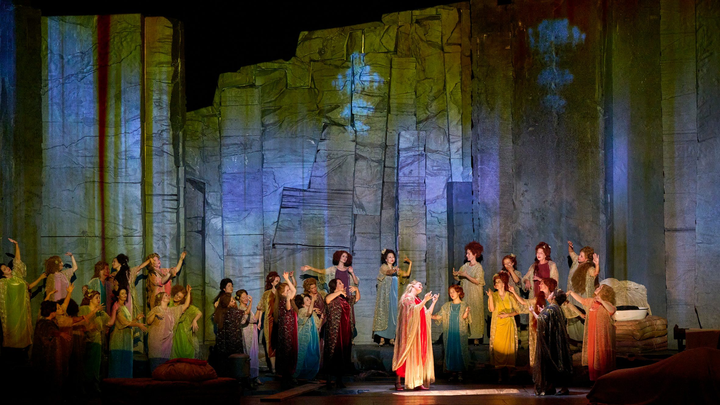 Eine Gruppe von Menschen in bunten Kleidern steht auf der Bühne vor einer großen, farbenfroh beleuchteten Felswand.