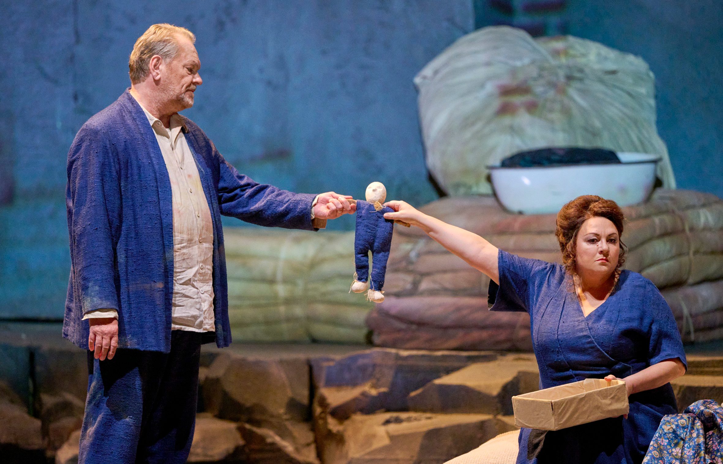 Zwei Opernsänger in blauen Kostümen auf der Bühne, eine Frau kniet und reicht einem Mann eine kleine Puppe.
