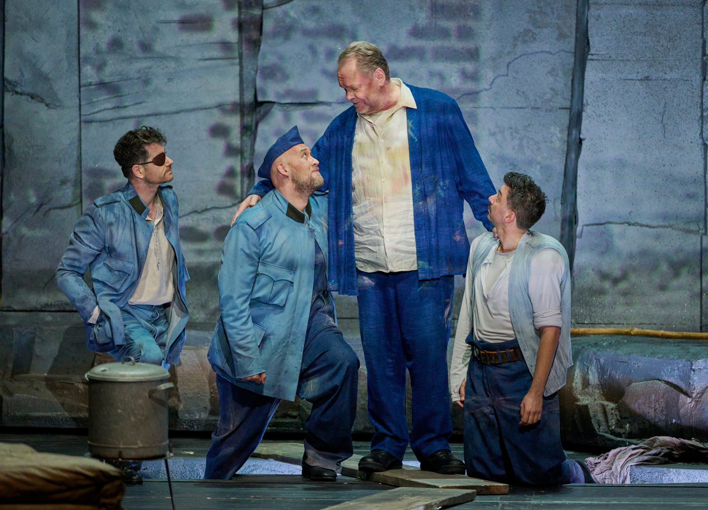 Vier Männer in blauen Kostümen stehen und knien auf der Bühne, einer legt dem Größten die Hand auf die Schulter.