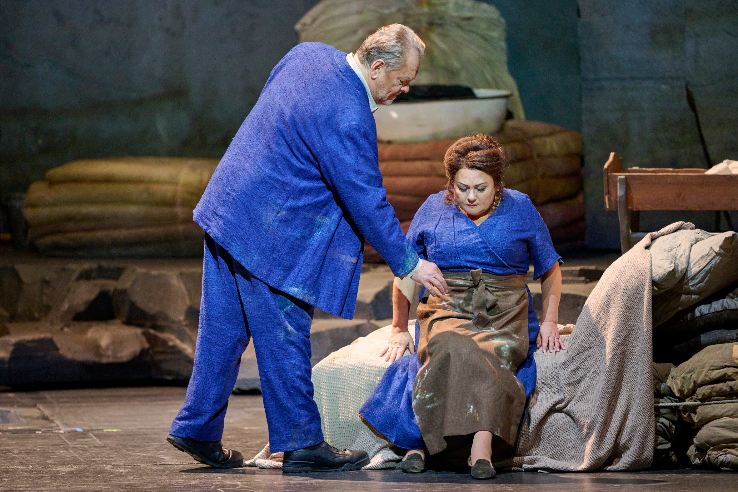 Ein Mann in blauem Kostüm beugt sich zu einer sitzenden Frau, die betroffen nach unten blickt.