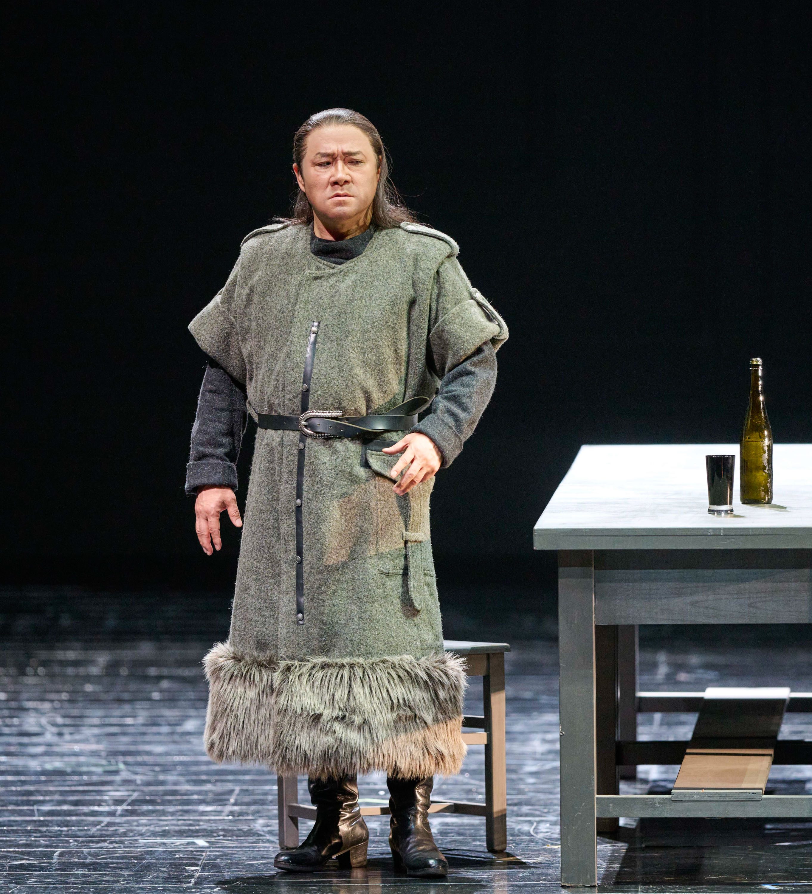 Ein Mann in mittelalterlicher Gewandung steht auf einer Bühne neben einem Tisch mit einer Flasche und einem Becher.