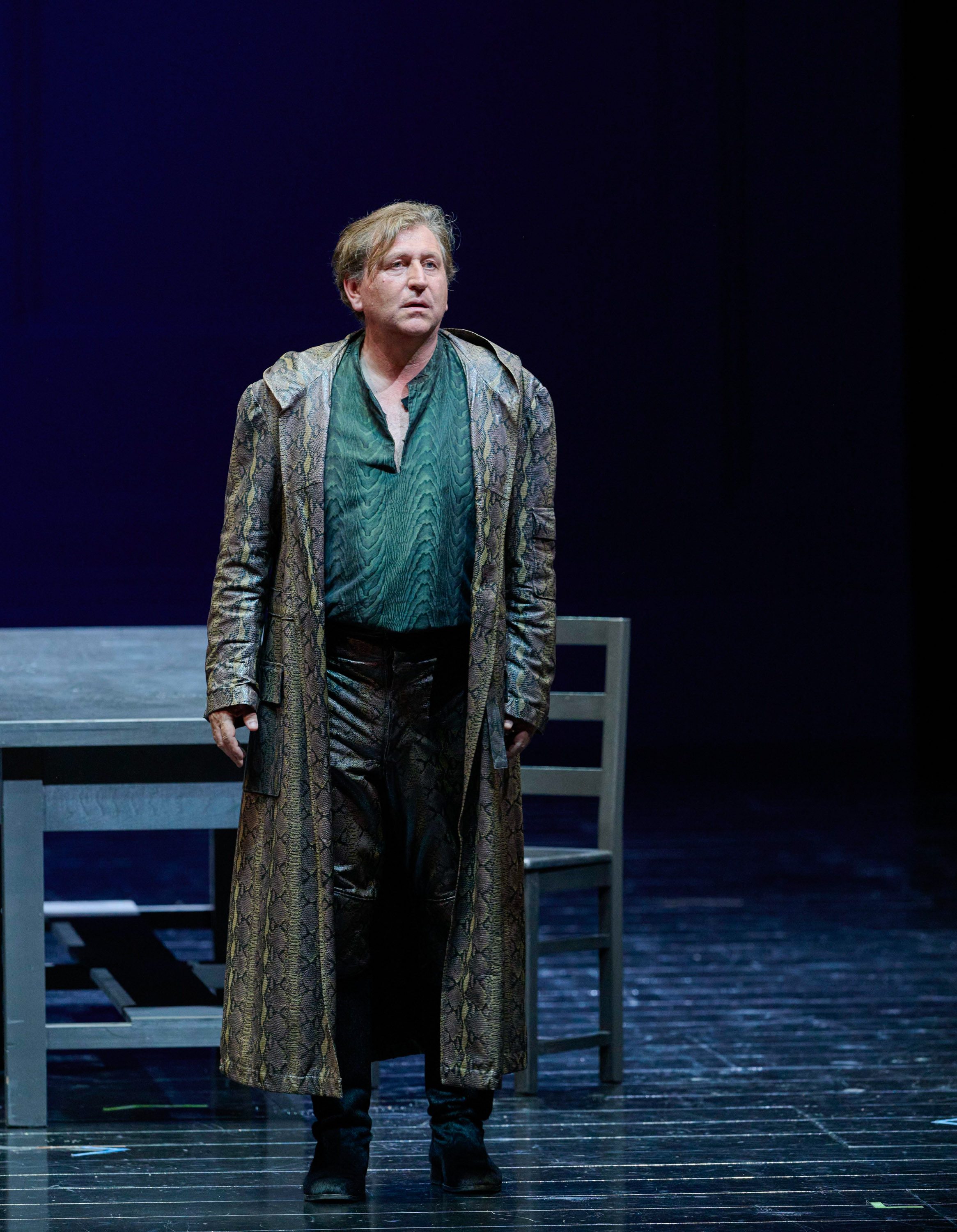 Ein Mann in langem Mantel und halb geöffnetem Hemd steht auf einer Bühne neben einem Tisch und Stuhl.