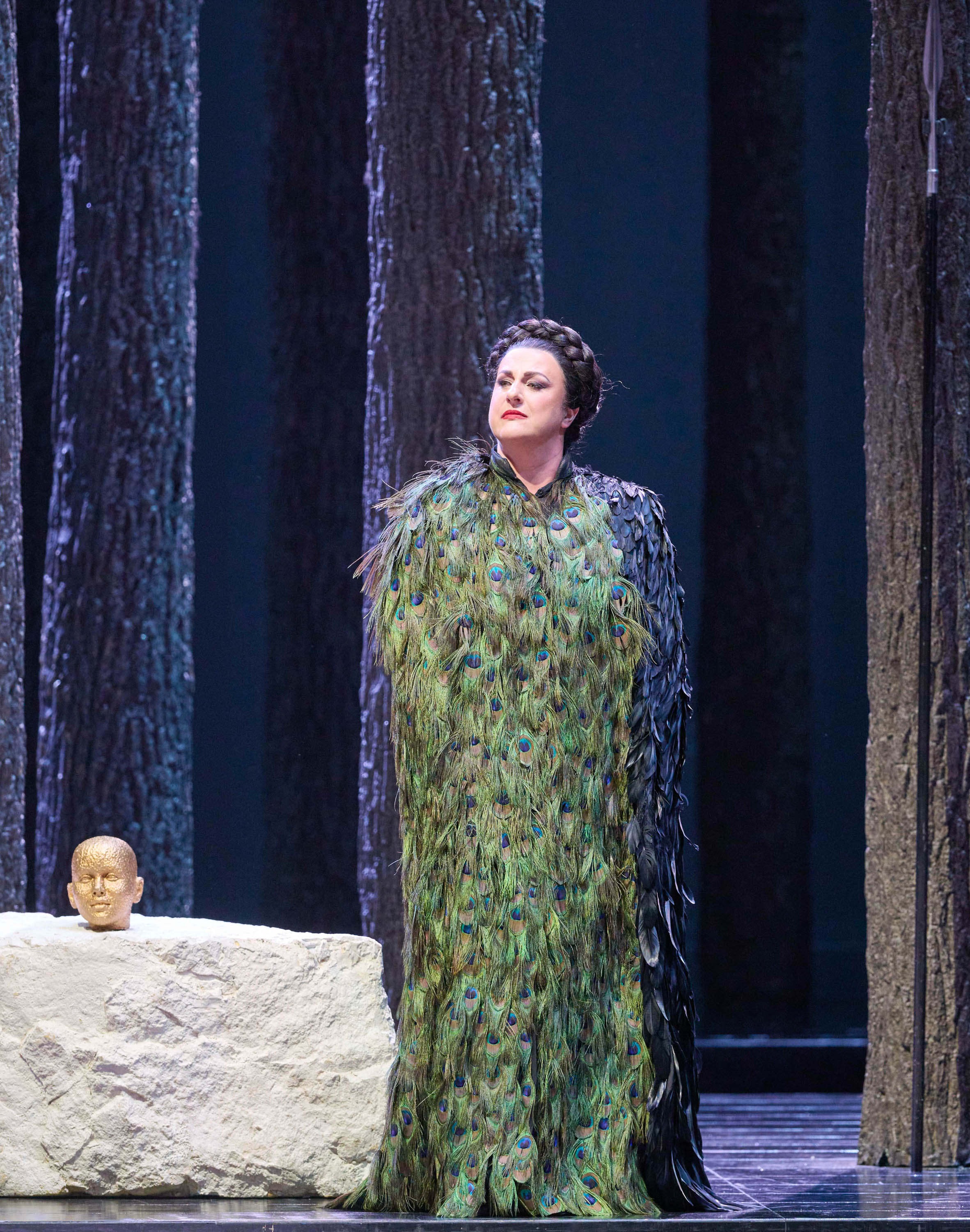 Eine Frau in grünem Federkleid steht auf einer Bühne vor einer Steinplatte mit einem kleinen Kopf darauf.