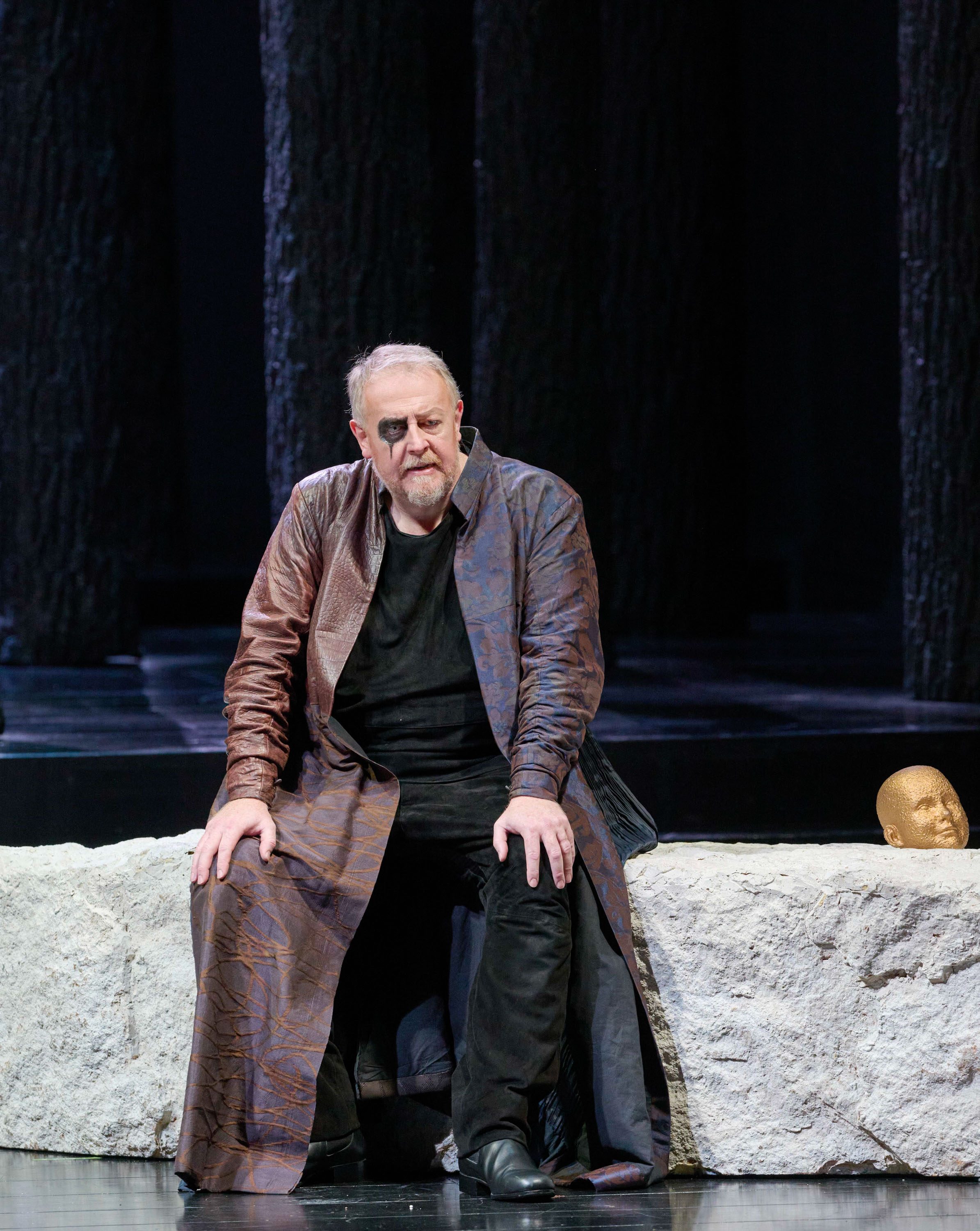 Ein Mann in langem Ledermantel sitzt auf einer Steinmauer mit einer Skulptur eines Kopfes daneben.