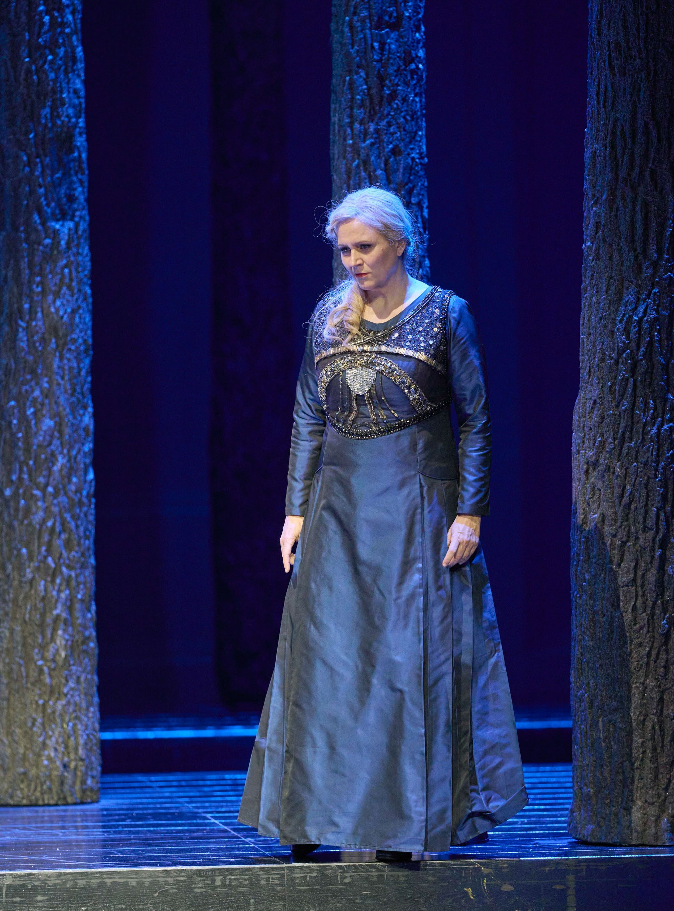 Eine Frau mit langen, geflochtenen Haaren und einem eleganten, blauen Kleid steht zwischen Bäumen auf einer Theaterbühne.