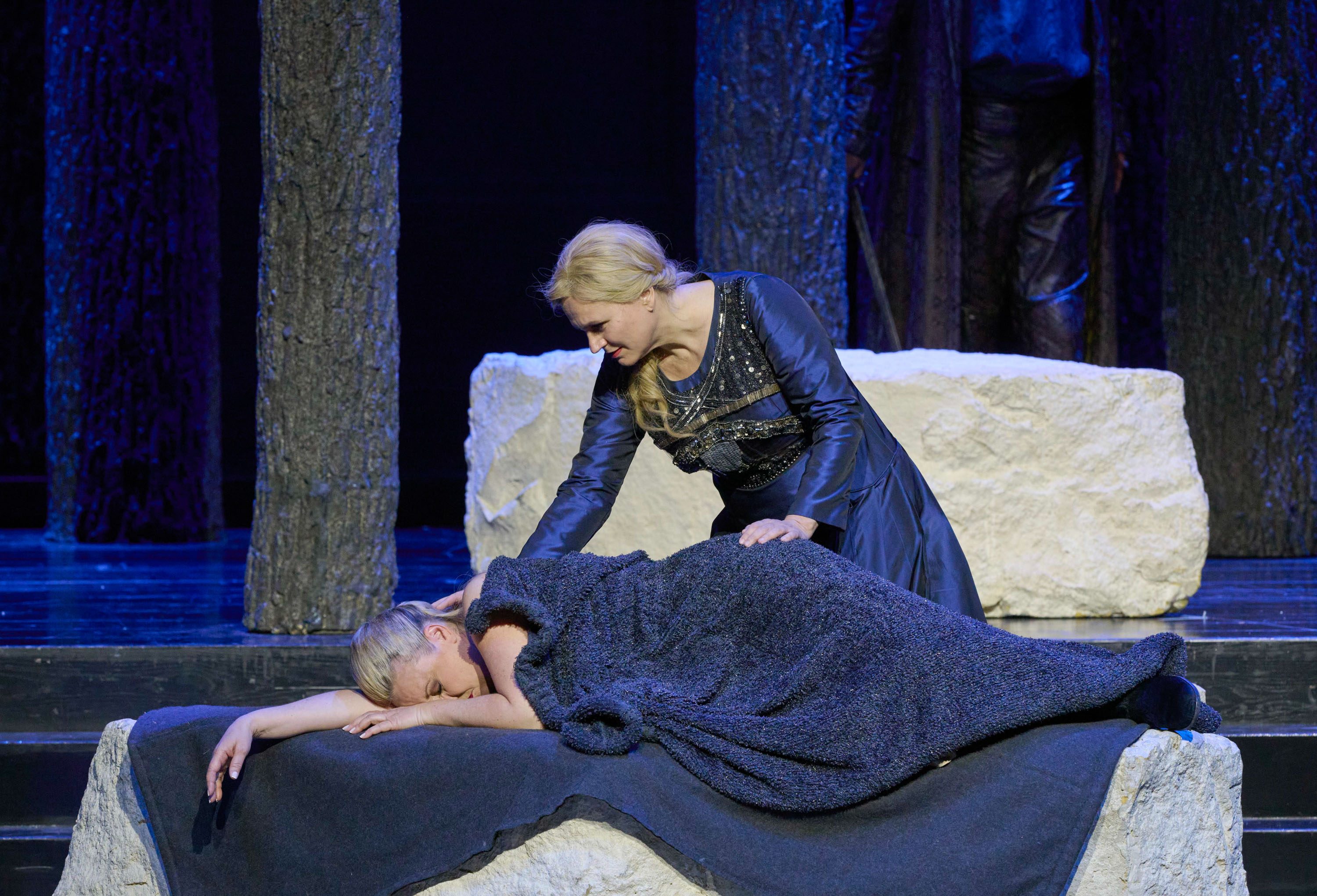 Eine blonde Frau in einem langen blauen Kleid streichelt liebevoll den Kopf einer schlafenden Frau, die in eine blaue Decke gehüllt ist.