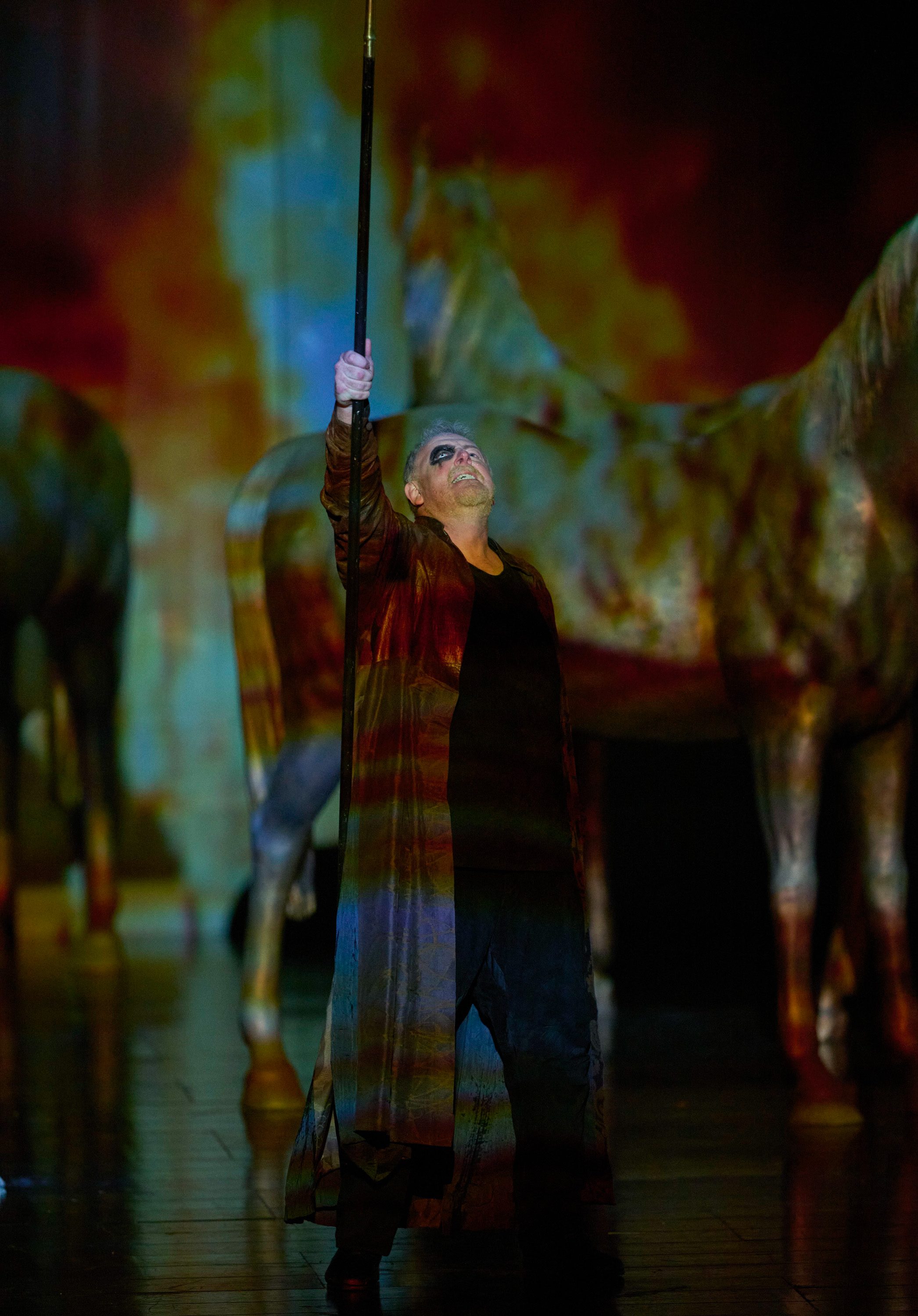 Eine Figur in langem Mantel hält eine Stange in die Luft, während im Hintergrund riesige Pferde zu sehen sind.