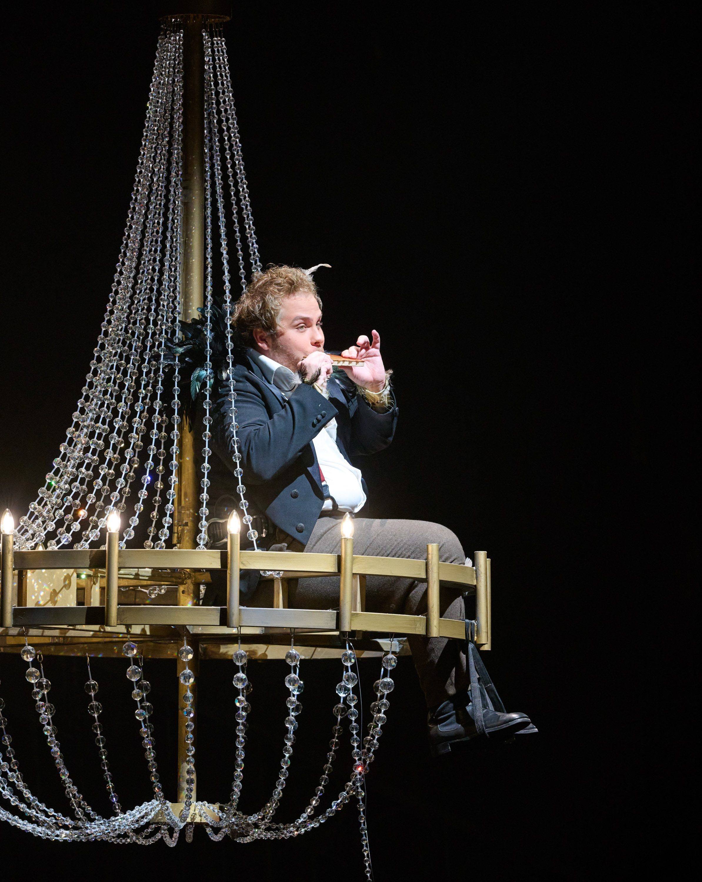 Ein Mann mit Federn in den Haaren, sitzt auf einem Kronleuchter und spielt Panflöte