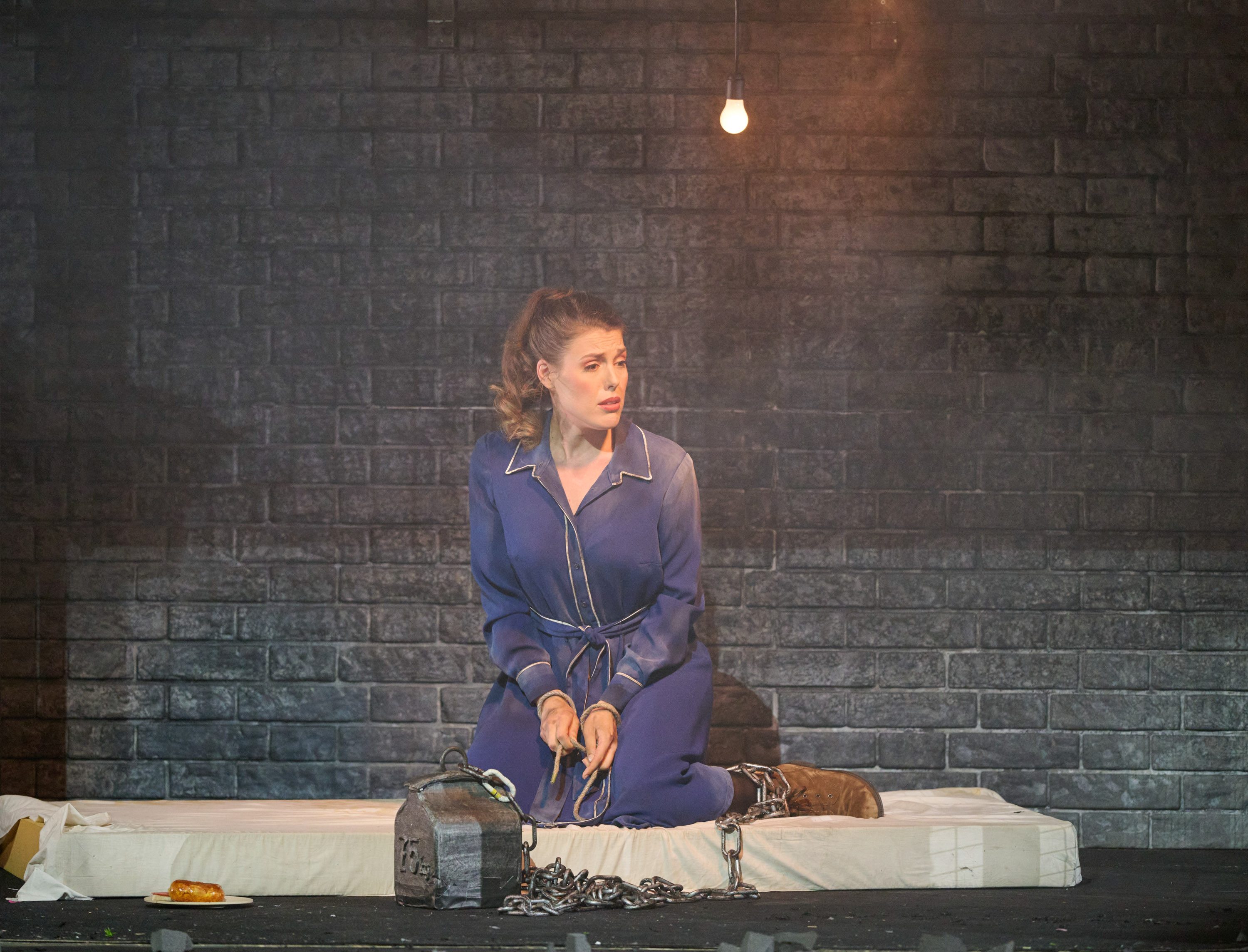 Eine Frau in einem schlichten blauen Kleid sitzt in Ketten gefesselt auf einer Matratze.