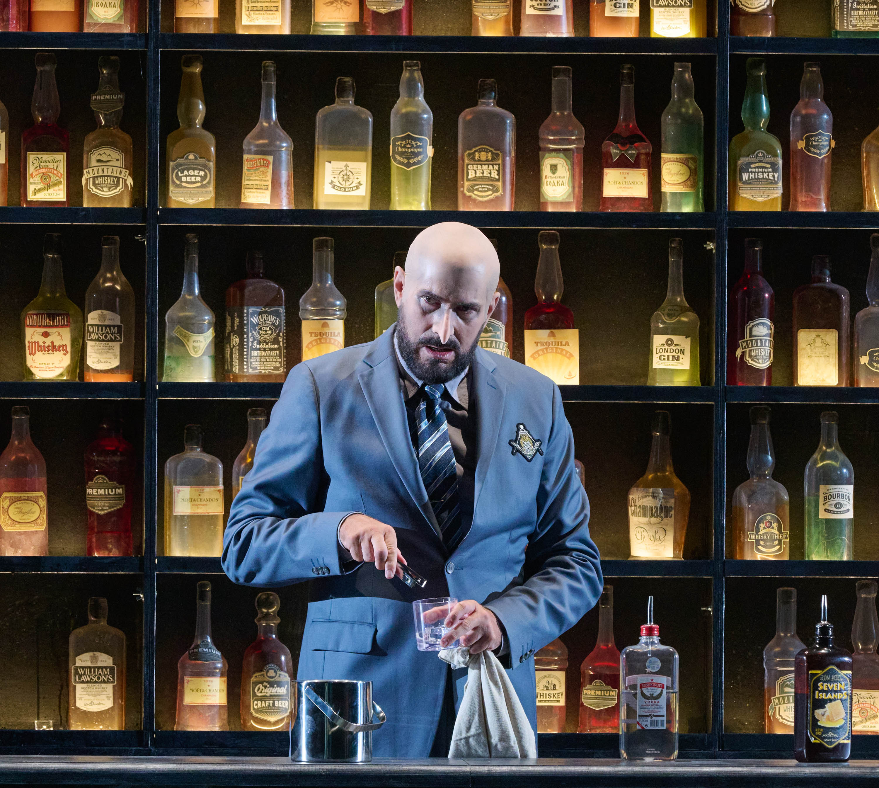 Ein Mann mit Glatze und Hakennase steht vor einer Bar und gießt etwas in ein Glas,