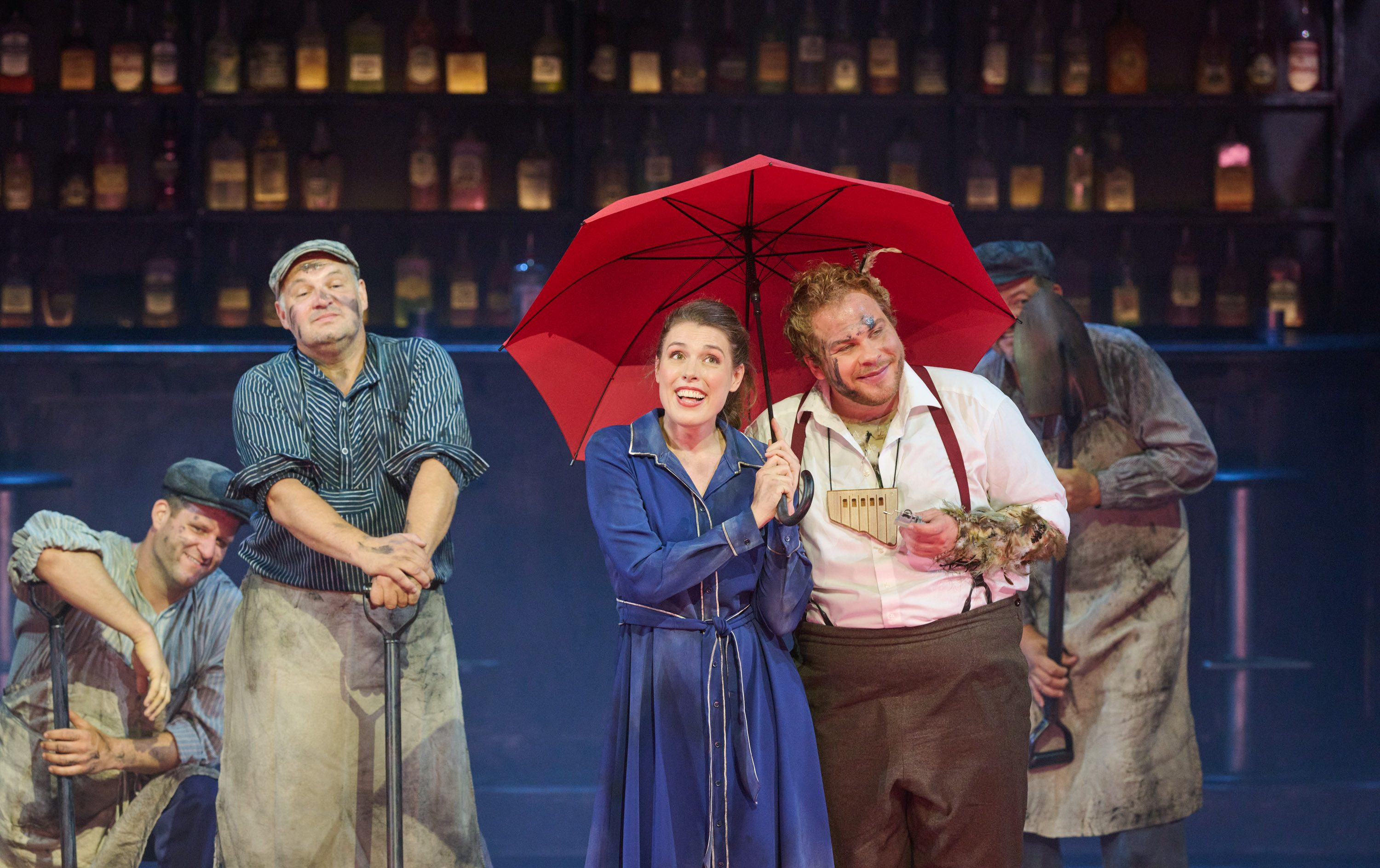 Ein Mann, der ein Federkostüm anhat und eine Panflöte um den Hals trägt steht mit einer Frau in einem blauen Kleid unter einem roten Regenschirm. Um sie herum stehen Männer, die mit Kohle verschmiert sind und Schaufeln in der Hand halten.