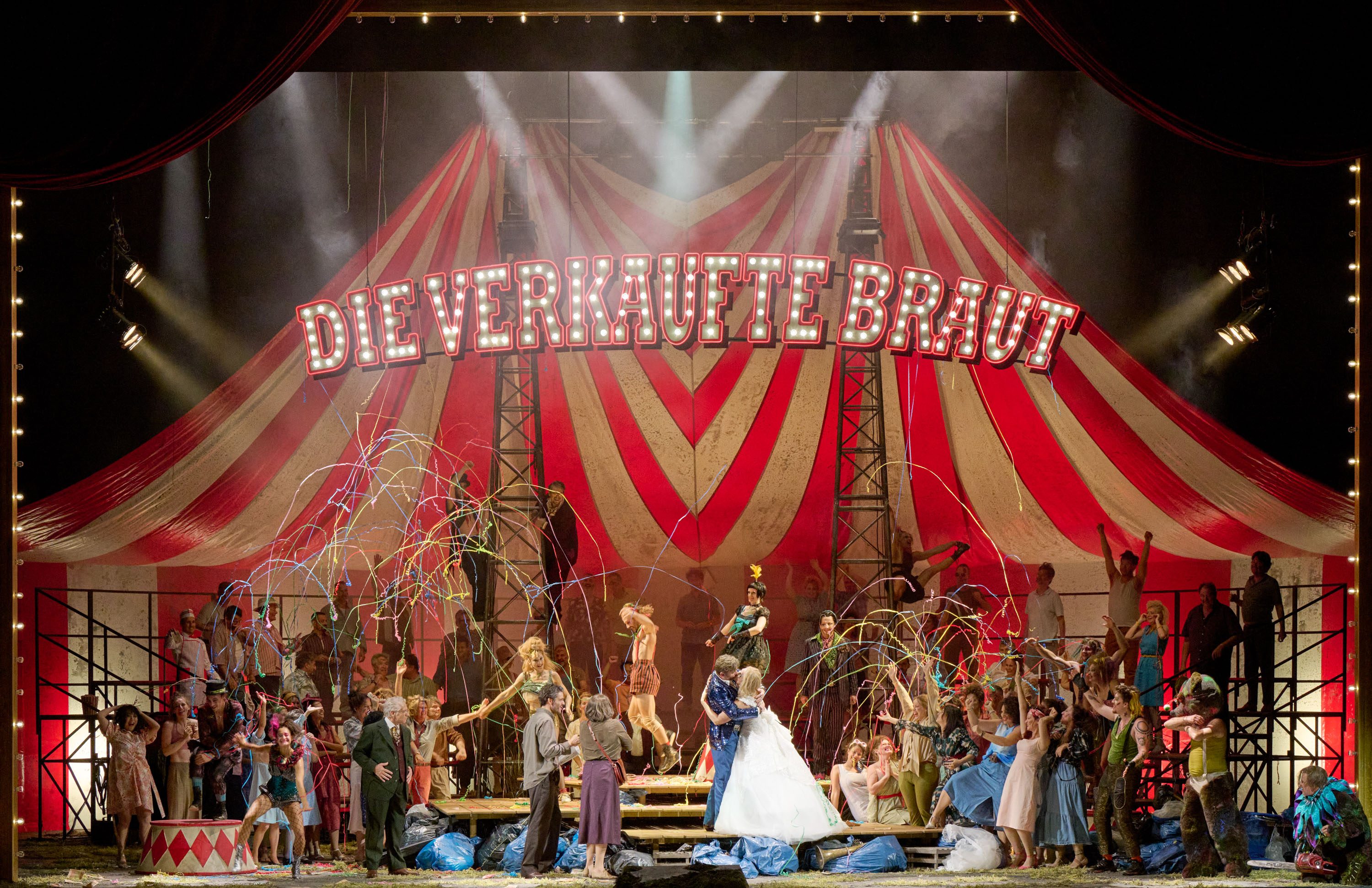 A vibrant theatrical scene from "Die verkaufte Braut," featuring performers celebrating on stage beneath a large striped tent. Colorful ribbons are being sprayed, and a couple in wedding attire is central to the celebration. Numerous characters are joyfully interacting, creating a festive atmosphere.