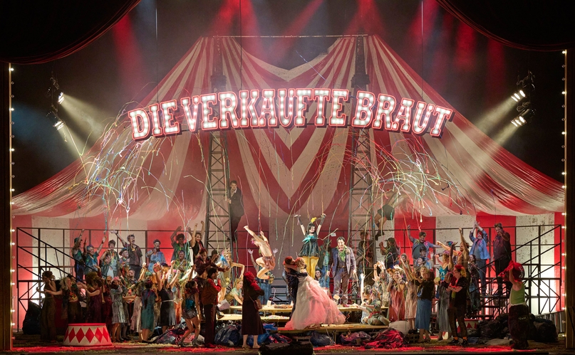 In einem großen Zirkuszelt, an dessen Decke der Schriftzug "Die verkaufte Braut" Hängt, sitzen viele bunt gekleidete Menschen. In der Mitte stehen ein Mann und eine Frau, die sich innig küssen. Von der Decke kommen Luftschlangen.