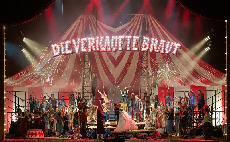 In einem großen Zirkuszelt, an dessen Decke der Schriftzug "Die verkaufte Braut" Hängt, sitzen viele bunt gekleidete Menschen. In der Mitte stehen ein Mann und eine Frau, die sich innig küssen. Von der Decke kommen Luftschlangen.