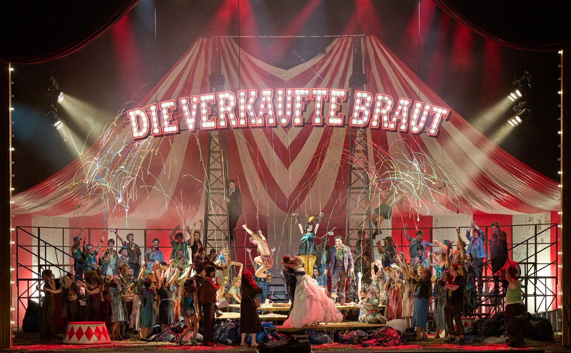 In einem großen Zirkuszelt, an dessen Decke der Schriftzug "Die verkaufte Braut" Hängt, sitzen viele bunt gekleidete Menschen. In der Mitte stehen ein Mann und eine Frau, die sich innig küssen. Von der Decke kommen Luftschlangen.