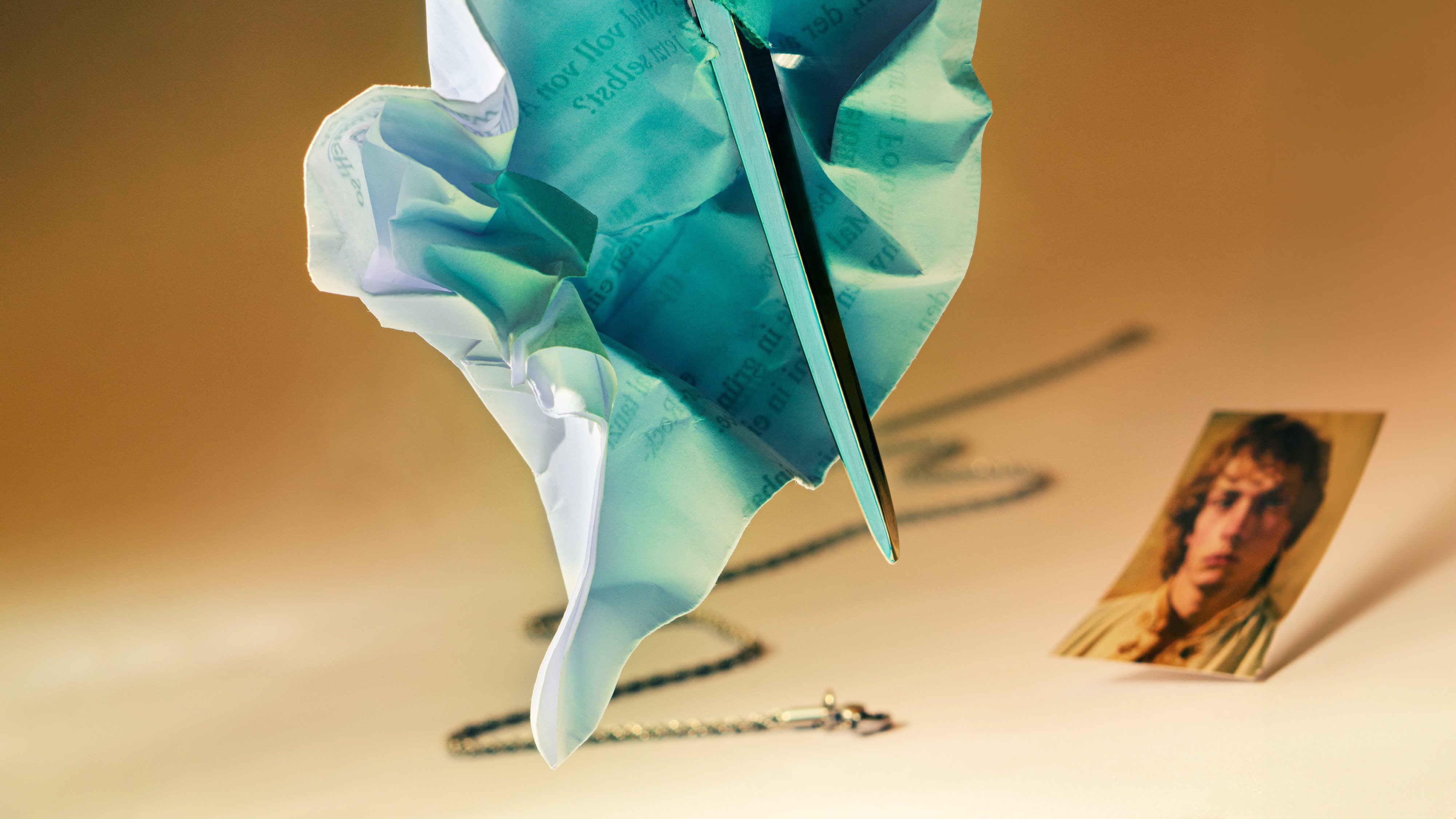 Ein türkisfarbenes Papier wird von einem Stab durchbohrt; eine goldene Kette und ein Foto liegen am Boden.