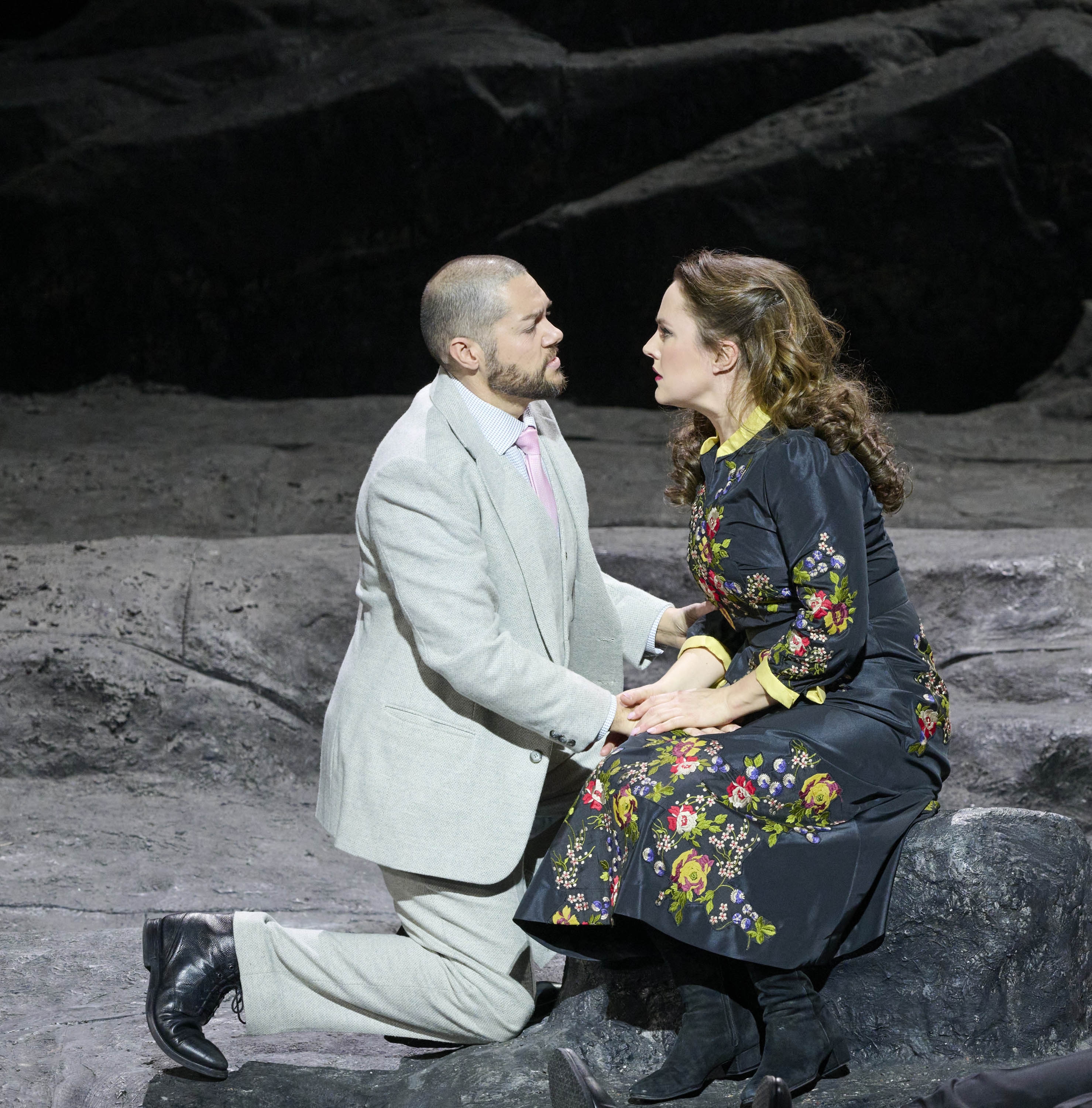 Ein Mann im grauen Anzug kniet vor einer Frau; sie befinden sich in einer Felsenlandschaft.