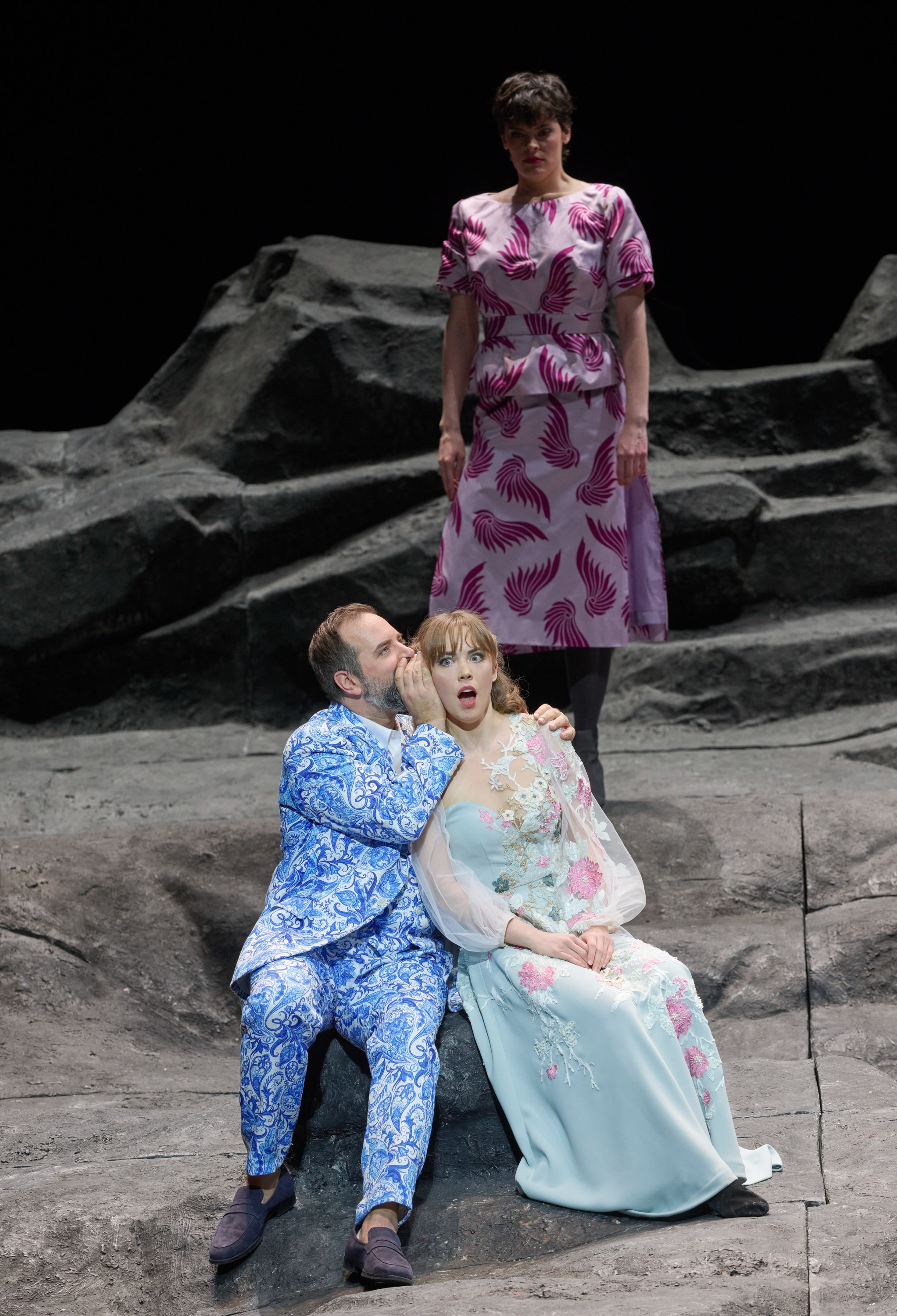 Ein Mann flüstert etwas in das Ohr einer Frau, die überrascht wirkt; im Hintergrund werden sie von einer anderen Frau beobachtet.