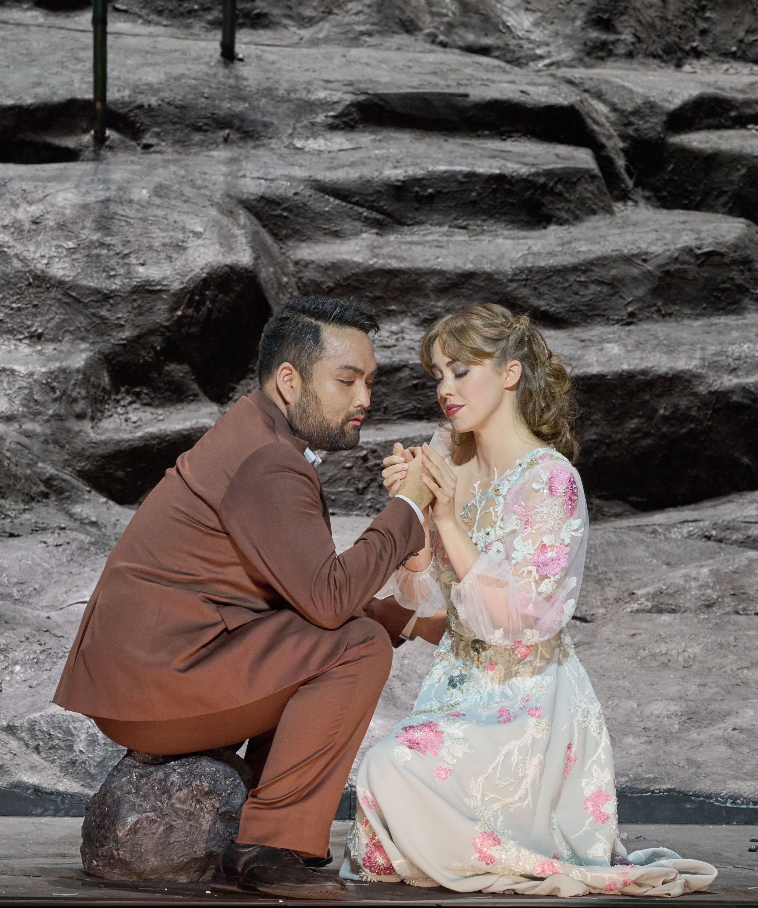 Eine Frau hält die Hand eines Mannes in braunem Anzug; sie befinden sich in einer grauen Felsenlandschaft.