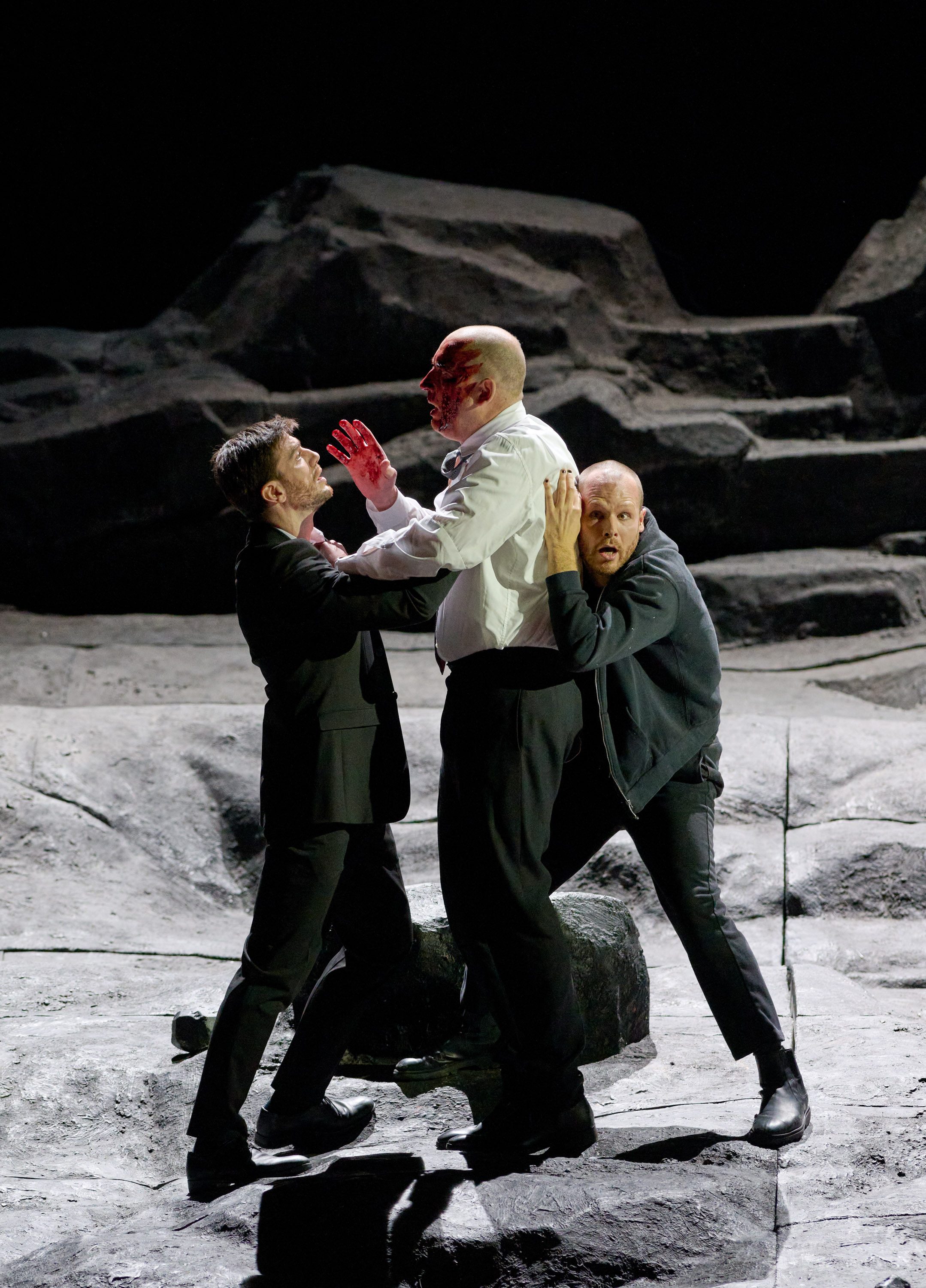 Drei Männer stehen auf einer steinigen Bühne. Der Mann in der Mitte trägt ein blutiges Hemd und schaut verzweifelt. Die anderen beiden versuchen, ihn festzuhalten oder zu beruhigen. Die Szenerie vermittelt eine angespannte Konfrontation.