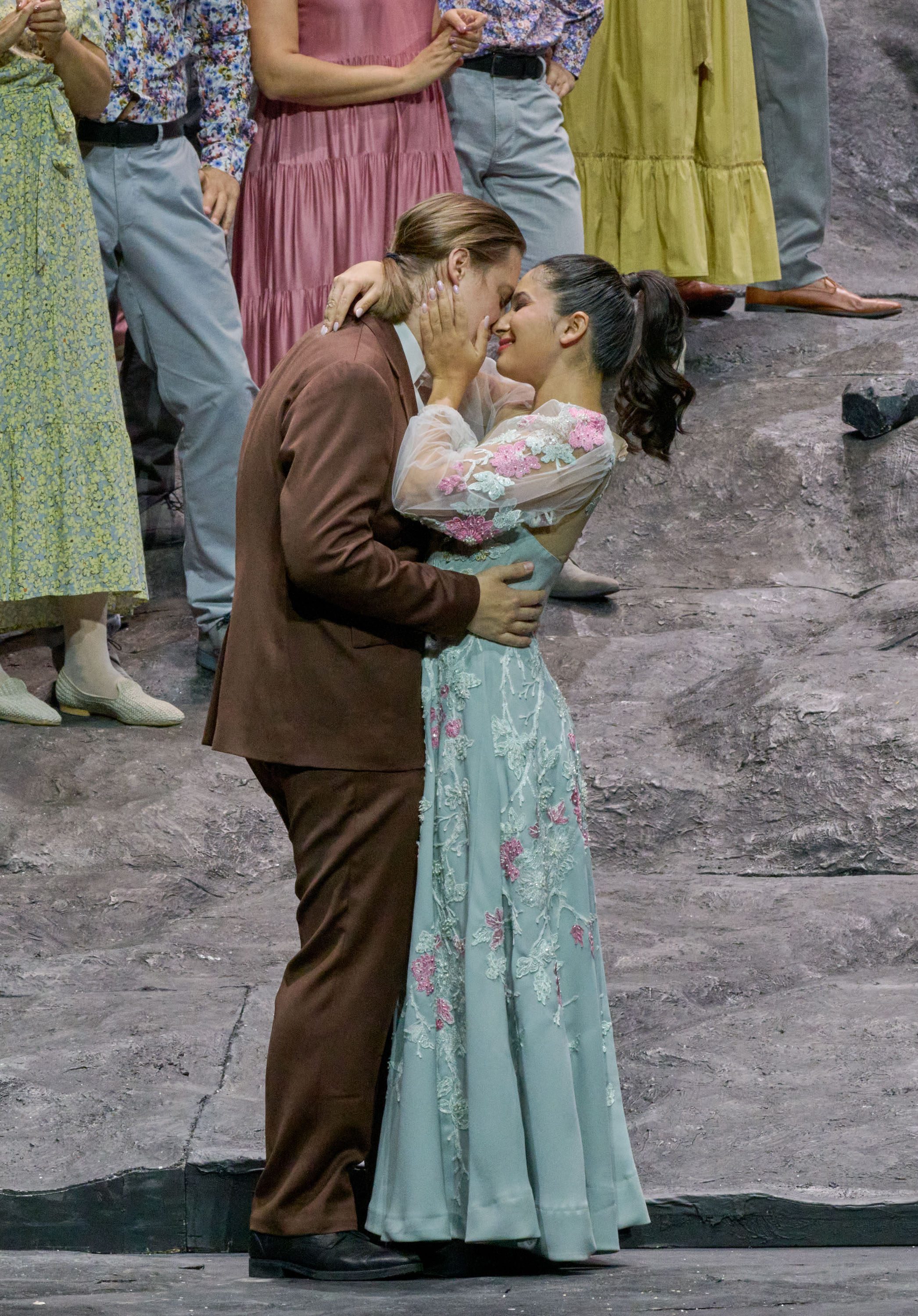 Ein verliebtes Paar steht eng umschlungen auf einer Bühnenkulisse. Sie schauen sich tief in die Augen und scheinen sich sanft zu küssen. Im Hintergrund sind weitere Personen in festlicher Kleidung zu sehen, die die intime Szene beobachten.