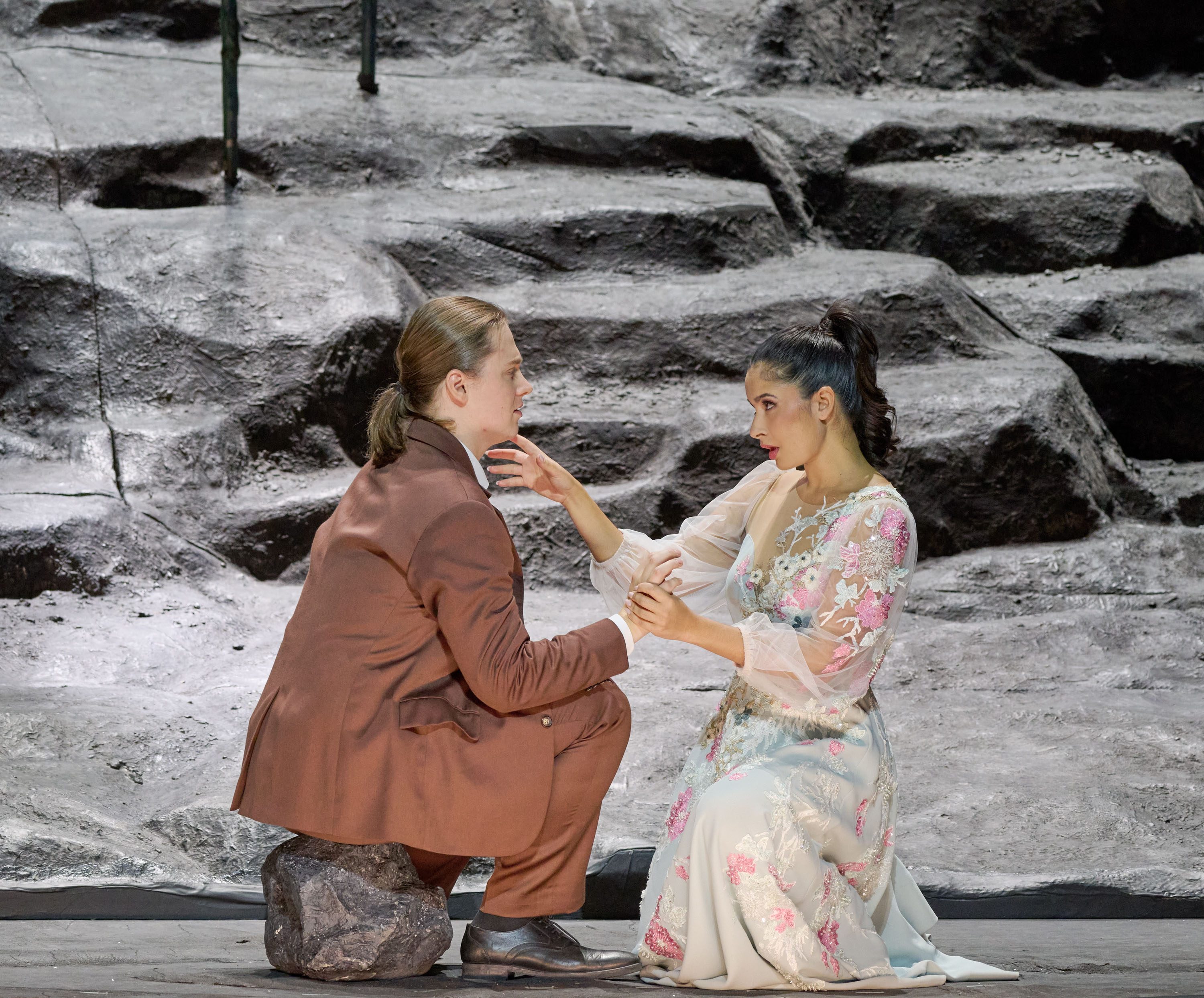 Zwei Darsteller auf einer Theaterbühne interagieren intensiv. Der männliche Darsteller kniet auf einem Stein und hält die Hand der weiblichen Darstellerin, die in einem aufwendigen, floral verzierten Kleid sitzt. Die Szene vermittelt Emotionen und Intimität zwischen den Charakteren.