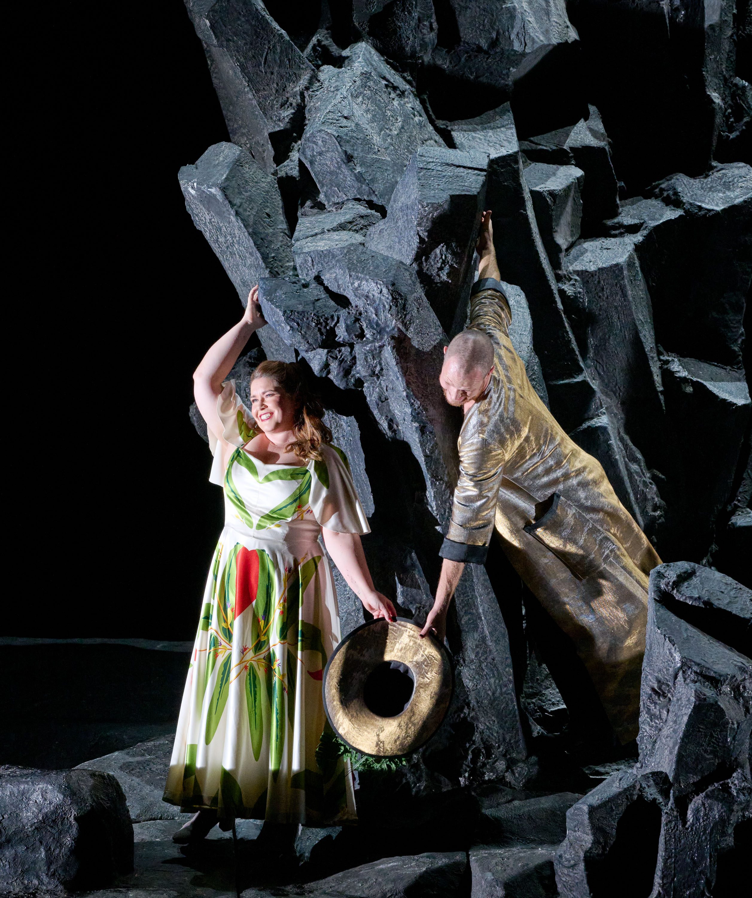 Eine Sängerin in einem blumenverzierten Kleid steht an einer Felswand und lächelt. Ein männlicher Darsteller in einem goldenen Outfit reicht ihr einen Hut. Die Szene vermittelt eine lebhafte, dramatische Atmosphäre.