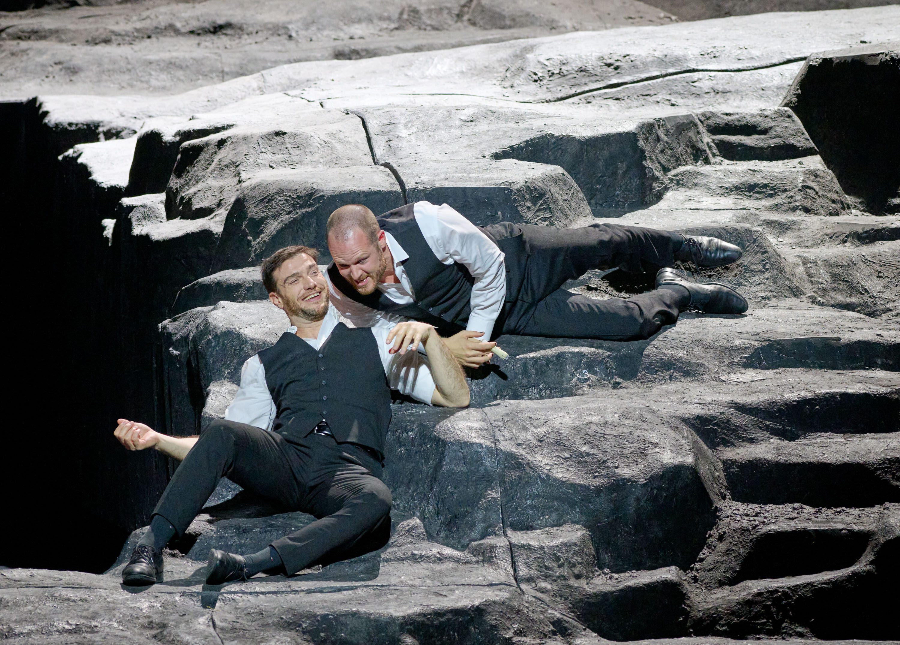Zwei Schauspieler liegen auf einer Felsoberfläche, lächeln und interagieren miteinander. Einer sitzt auf dem Boden, der andere beugt sich zu ihm über und greift nach seiner Hand. Beide tragen formelle Kleidung und scheinen sich in einer intensiven, aber freundlichen Situation zu befinden.