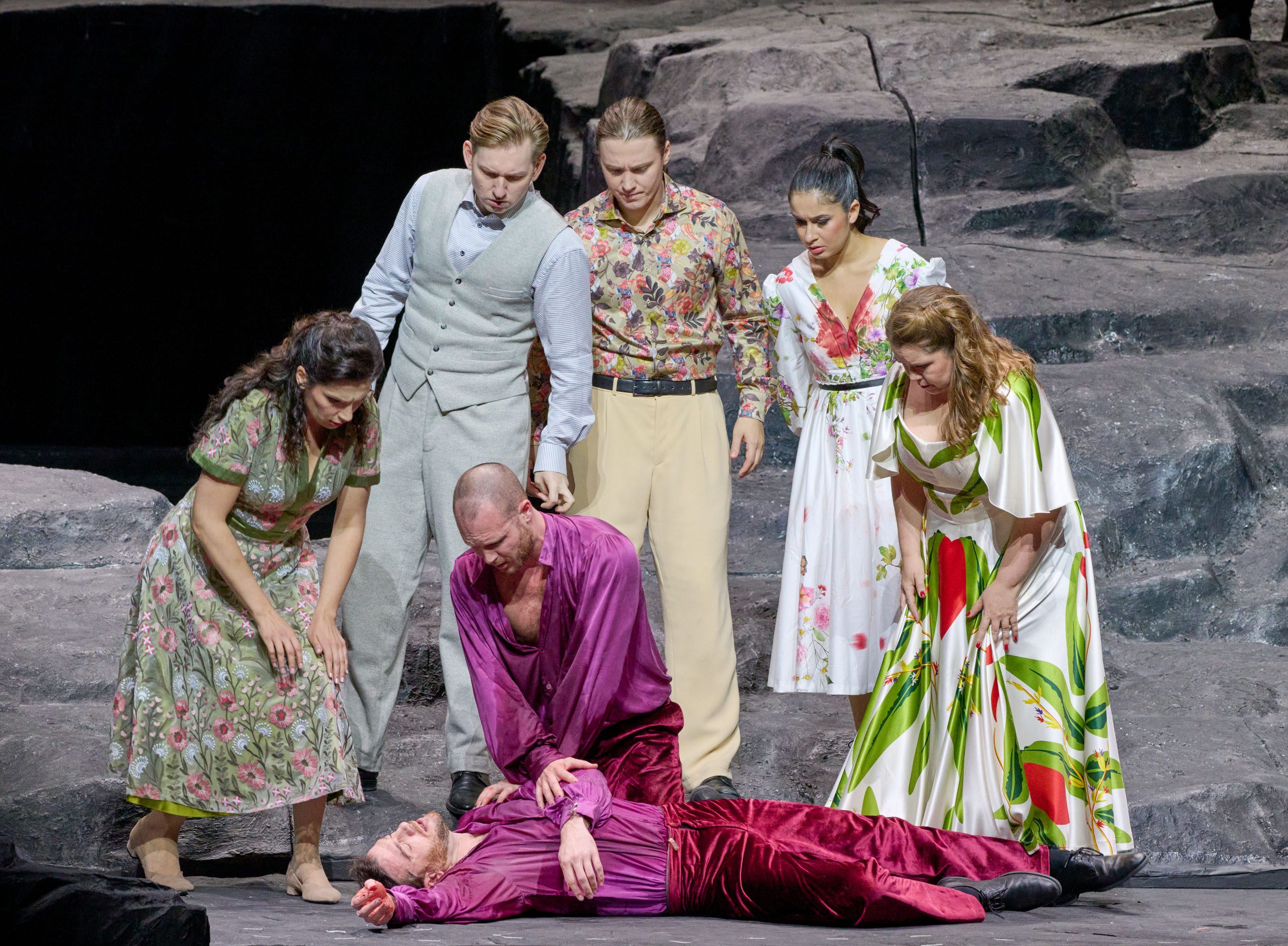Eine Gruppe von sechs Personen steht um einen Mann, der auf dem Boden liegt. Der Mann trägt ein lila Hemd und sieht verletzt aus. Die anderen schauen besorgt und versuchen ihm zu helfen. Die Frauen tragen farbenfrohe Kleider, die Männer formelle Kleidung. Der Hintergrund ist felsig.