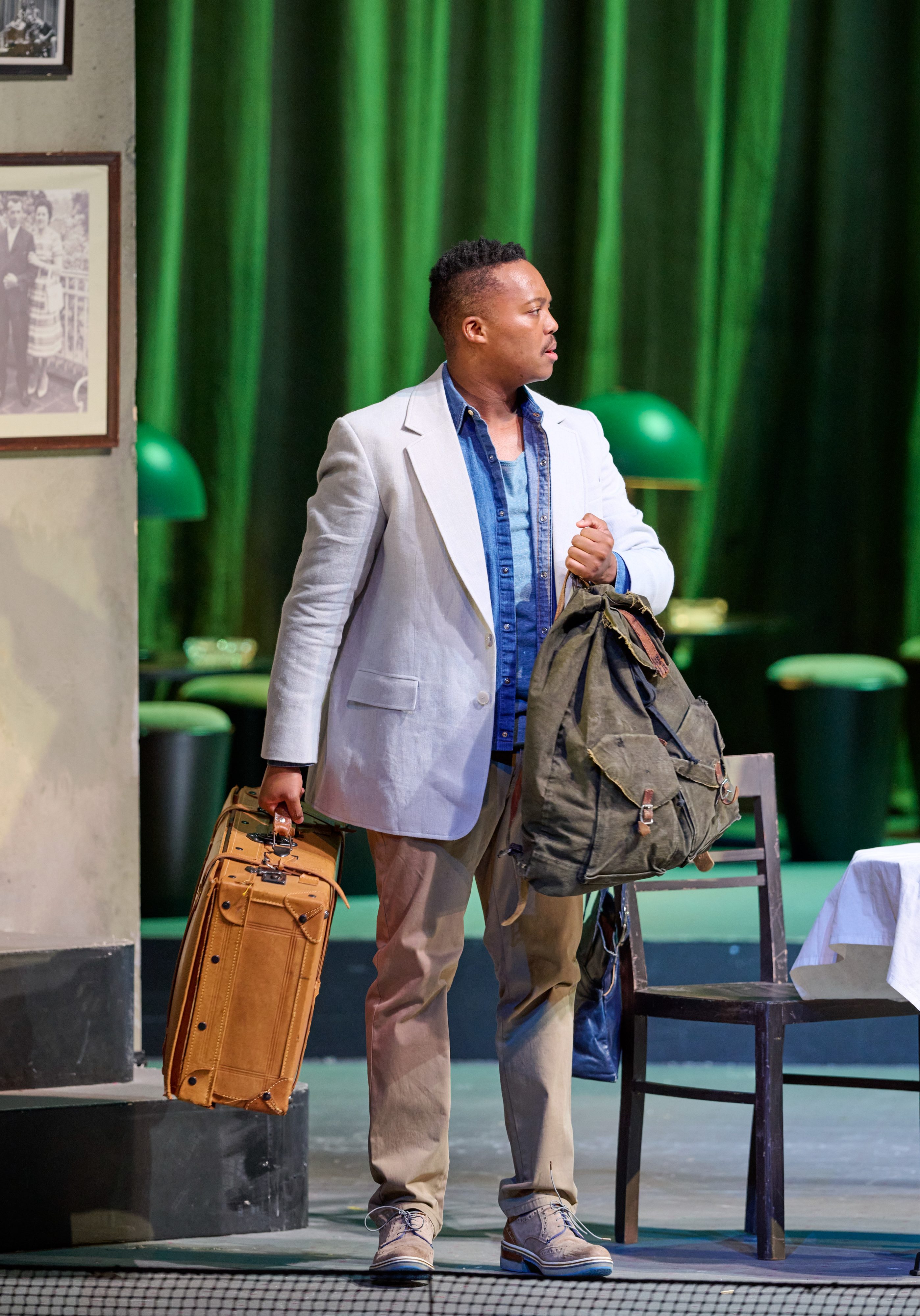 Ein Mann steht mit Gepäckstücken in der Hand auf einer Bühne vor einer Kulisse mit grünem Vorhang und Lampen.
