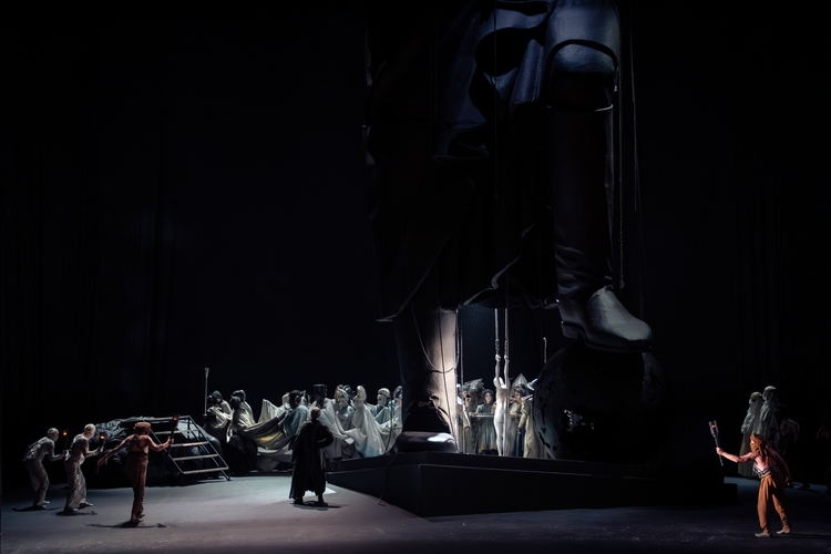 A dramatic stage scene features a large statue looming over performers. The front depicts a figure holding a torch, while others, draped in flowing costumes, appear to be interacting with the giant statue. The setting is dark, emphasizing the powerful contrast between light and shadow.