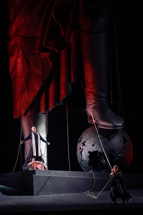 The image features a dramatic theatrical scene with a large statue-like figure in the background, illuminated in red. In the foreground, a person stands confidently on a stage, while another figure pulls on a rope, suggesting a struggle or tension. The atmosphere conveys a sense of conflict and power dynamics.