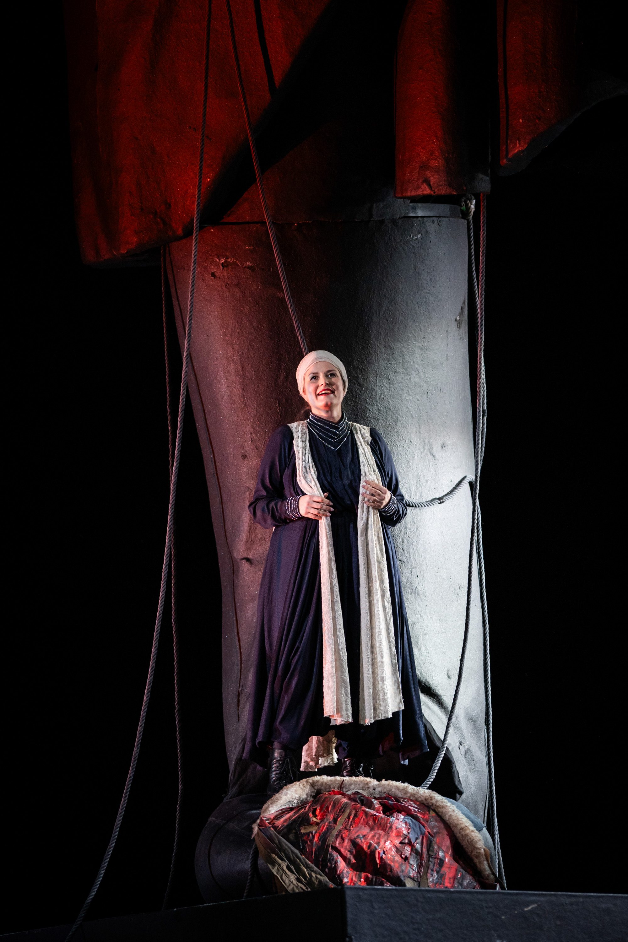 Eine Frau in einem langen, dunklen Kleid steht lächelnd vor einem großen, statueartigen Objekt. Sie trägt eine hellere Schürze und einen Kopftuch. Der Hintergrund ist dunkel, mit roten Lichtern, die eine dramatische Atmosphäre schaffen.