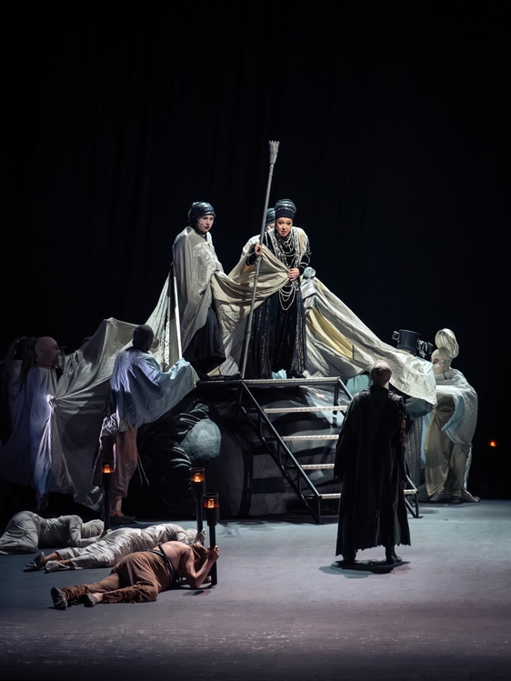 A dramatic theater scene features a central figure in dark attire holding a staff, surrounded by supporting characters draped in fabric. The backdrop is dark, and some figures appear to be fallen or subdued, creating an intense, somber atmosphere that suggests a narrative of struggle or conflict.