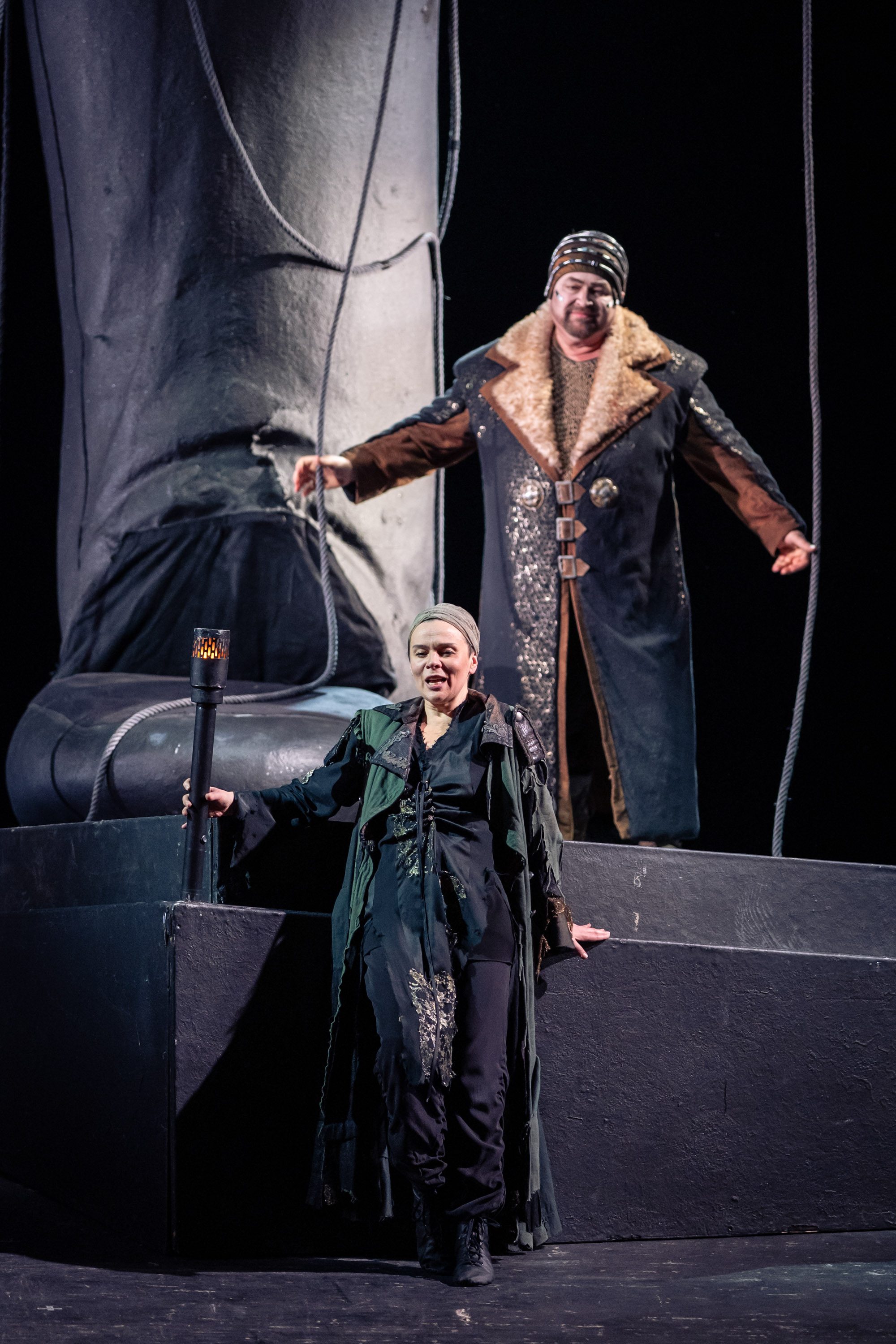 Eine Frau steht im Vordergrund und trägt einen langen, dunklen Mantel. Sie hält einen Stock in der Hand. Im Hintergrund steht ein Mann mit einem dicken Mantel und einem Hut. Beide scheinen Teil einer Theateraufführung zu sein, vor einer großen Statue oder einem Fuß.