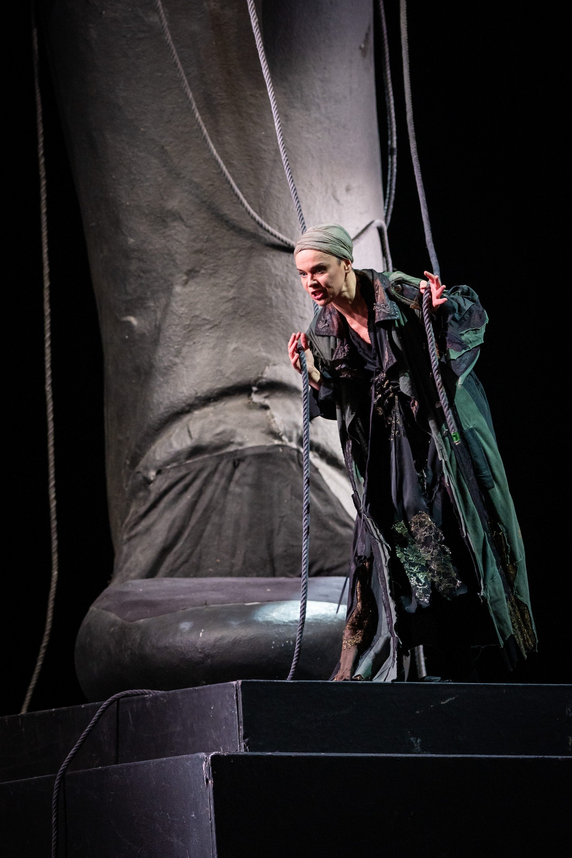 Eine schauspielende Person in einem langen, dunklen Gewand steht auf einer Bühne. Sie hält Seile in beiden Händen und schaut engagiert in die Ferne. Im Hintergrund ist ein großer, statuarischer Fuß sichtbar, der die Szene dominiert.