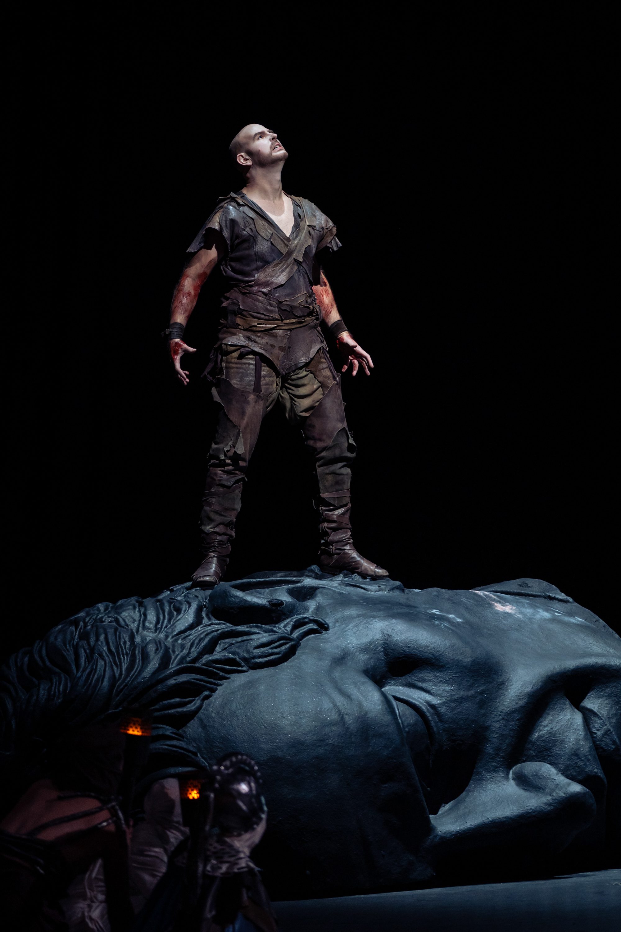 Ein männlicher Darsteller steht auf einem massiven Kopf, blickt nach oben und hat eine kraftvolle Pose eingenommen. Er trägt zerrissene, dreckige Kleidung und hat blutige Hände. Der Hintergrund ist dunkel, und die gesamte Szene vermittelt ein Gefühl von Dramatik und Intensität.