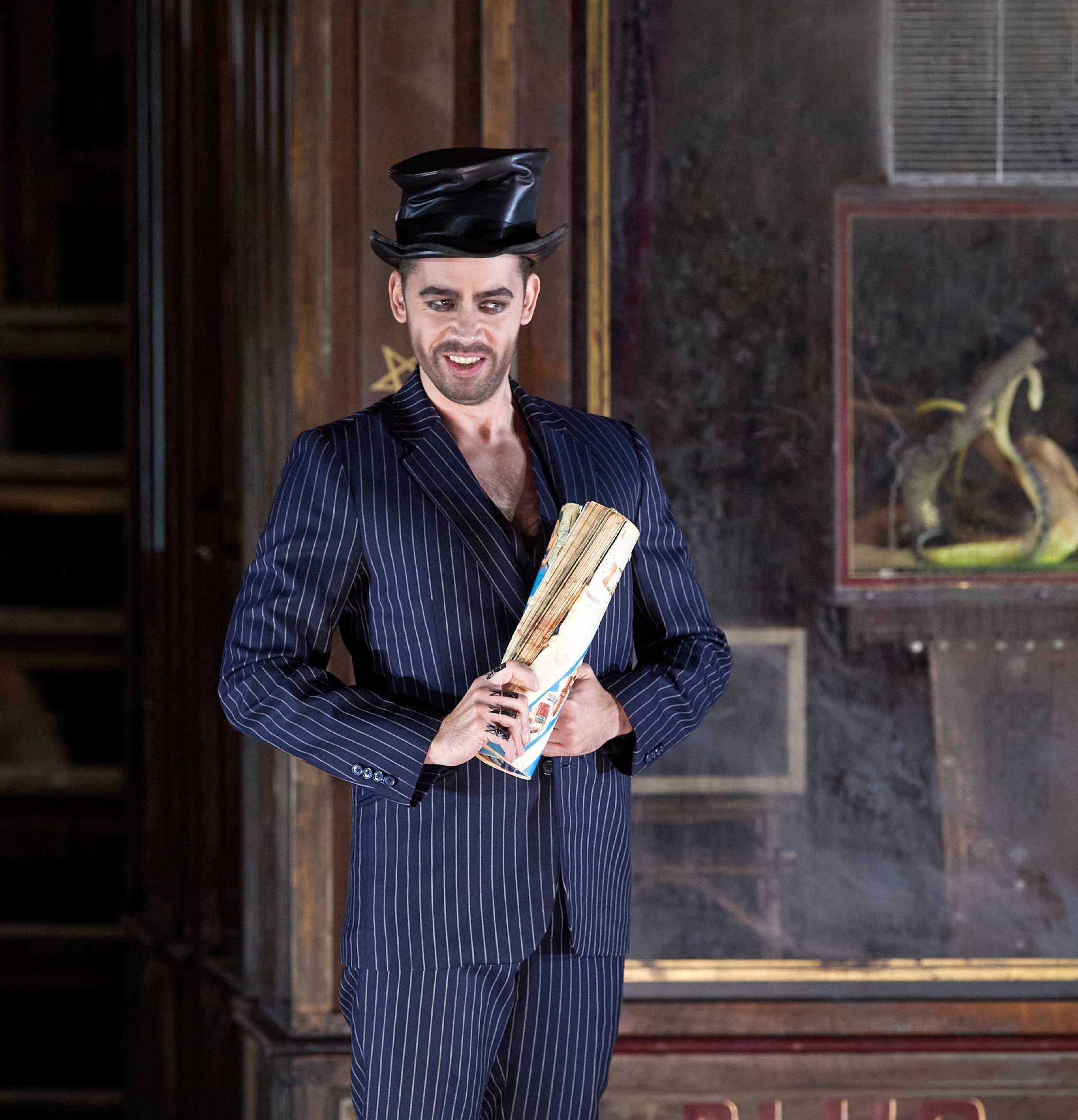 Ein Mann im gestreiften Anzug und glänzendem Zylinder hält lässig ein Fächerbündel in den Händen und blickt mit schelmischem Ausdruck.