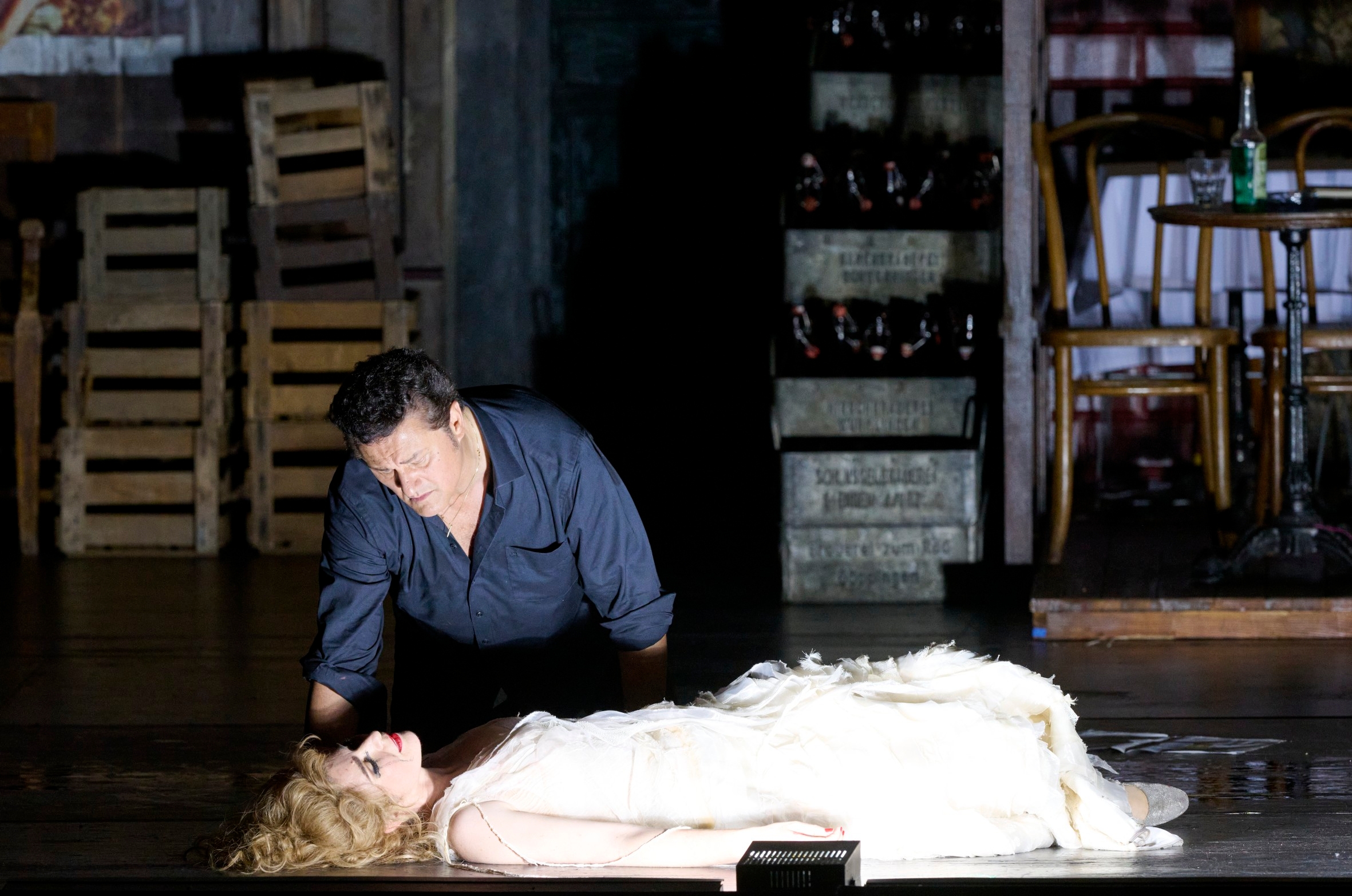 Ein Mann kniet verzweifelt neben einer regungslos auf dem Boden liegenden Frau im weißen Kleid auf einer düsteren Bühne.